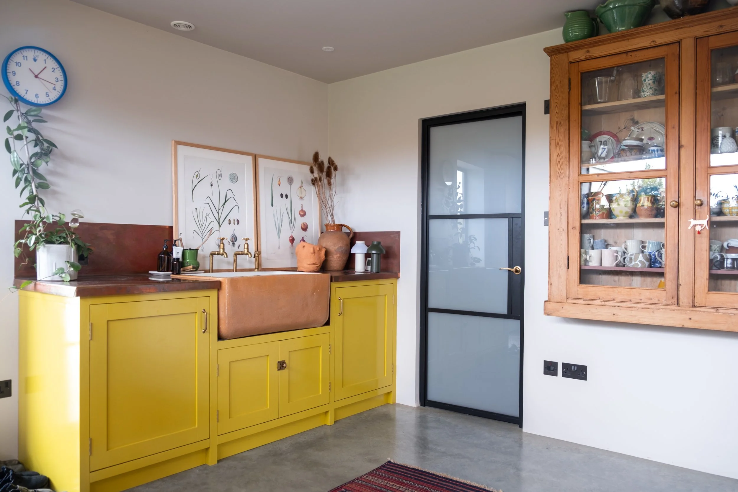 A kitchen corner with a yellow cabinet, a brown sink, and decorative items including framed botanical prints, pottery, and plants. There is a black-framed frosted glass door on the right and a wooden display cabinet filled with dishes and glassware o