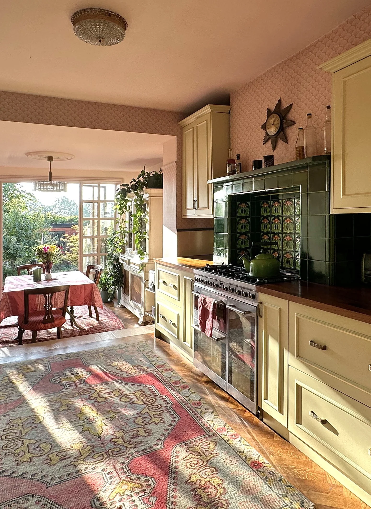 A cozy kitchen with yellow cabinets, a vintage green tiled fireplace, a wooden countertop, and a bright dining area with a table, chairs, and a view of greenery outside through large windows, with sunlight streaming in.