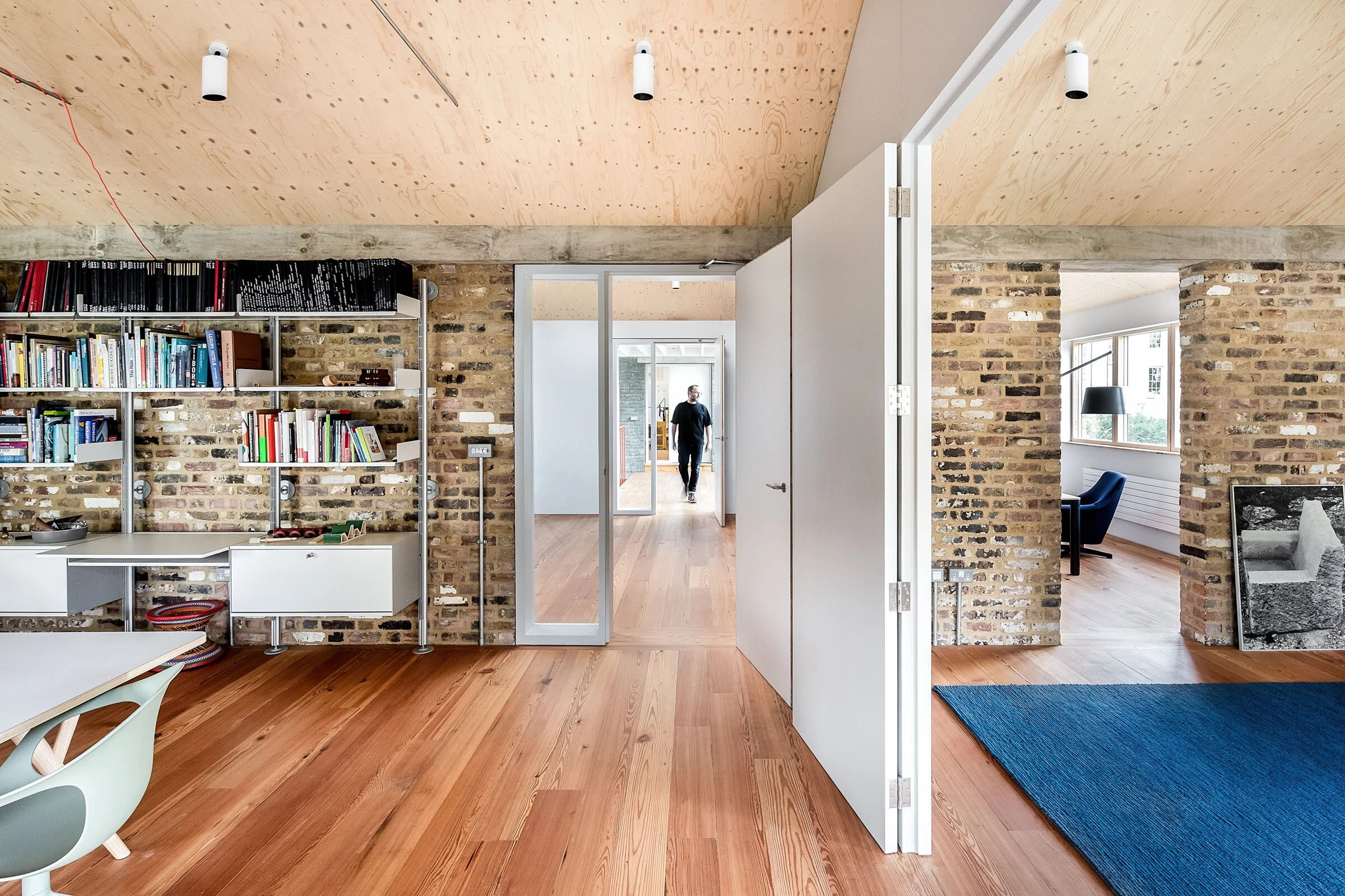Interior of a modern apartment with exposed brick walls and wooden floors, featuring a doorway leading to a hallway with a person walking, and rooms with bookshelves, large windows, and furniture.