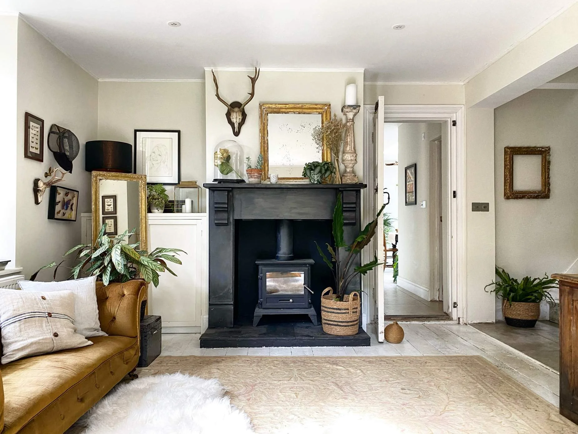 Living room with a yellow sofa, white pillows, a white fluffy rug, a black fireplace with potted plants on top and a large plant in a woven basket beside it, wall decor including framed pictures and a mounted deer skull with antlers, an open door lea