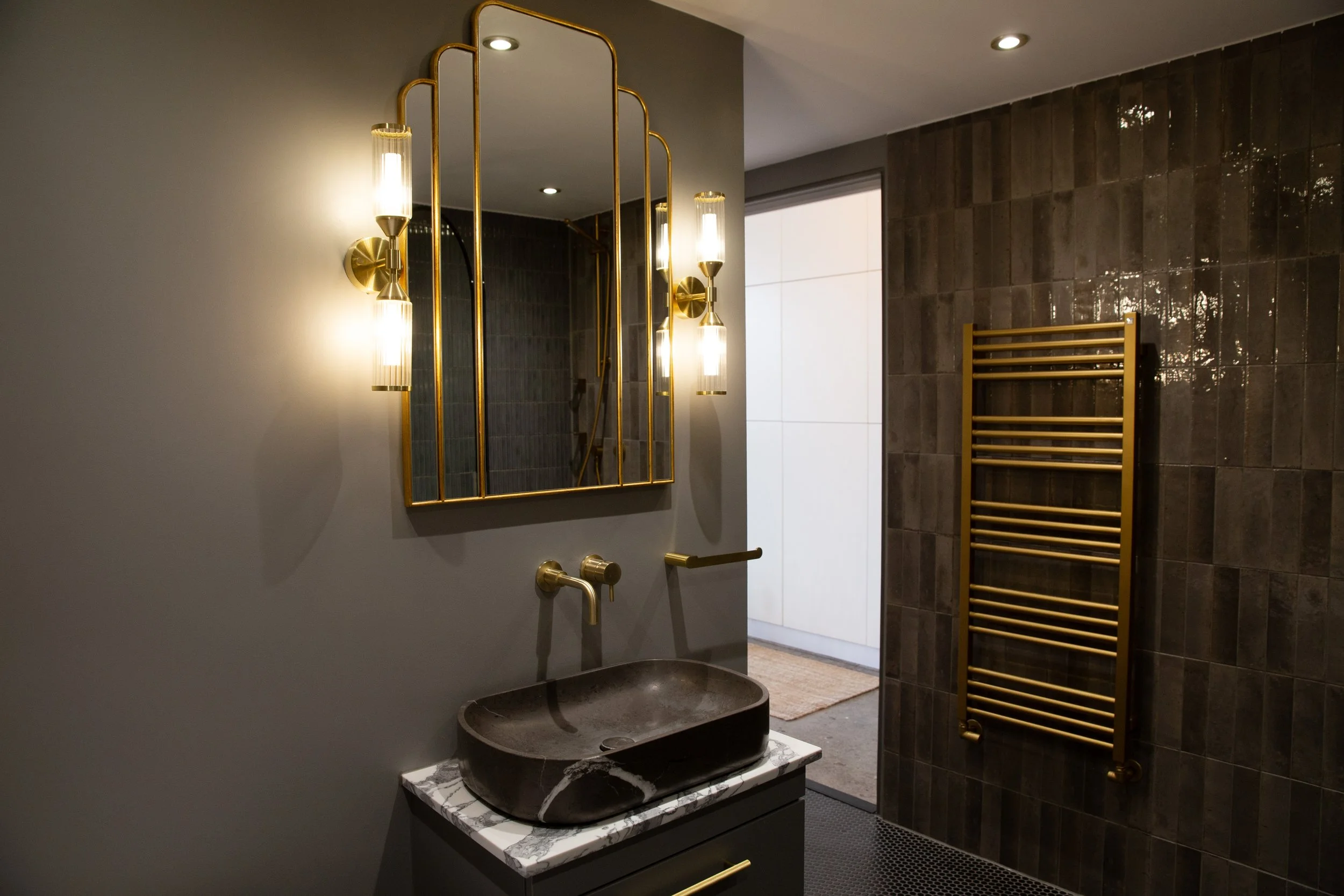 Modern bathroom featuring a black vessel sink on a marble countertop, gold fixtures, a decorative mirror with gold framing, wall-mounted wall sconces, and a heated towel rack on dark tiled wall.