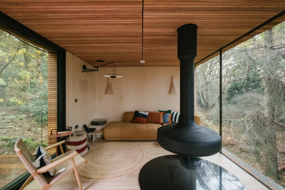 Modern living room with large glass windows showing trees, a beige sofa with multicolored pillows, a woven rug, a wood ceiling, a black fireplace, and a wooden chair with a pillow.