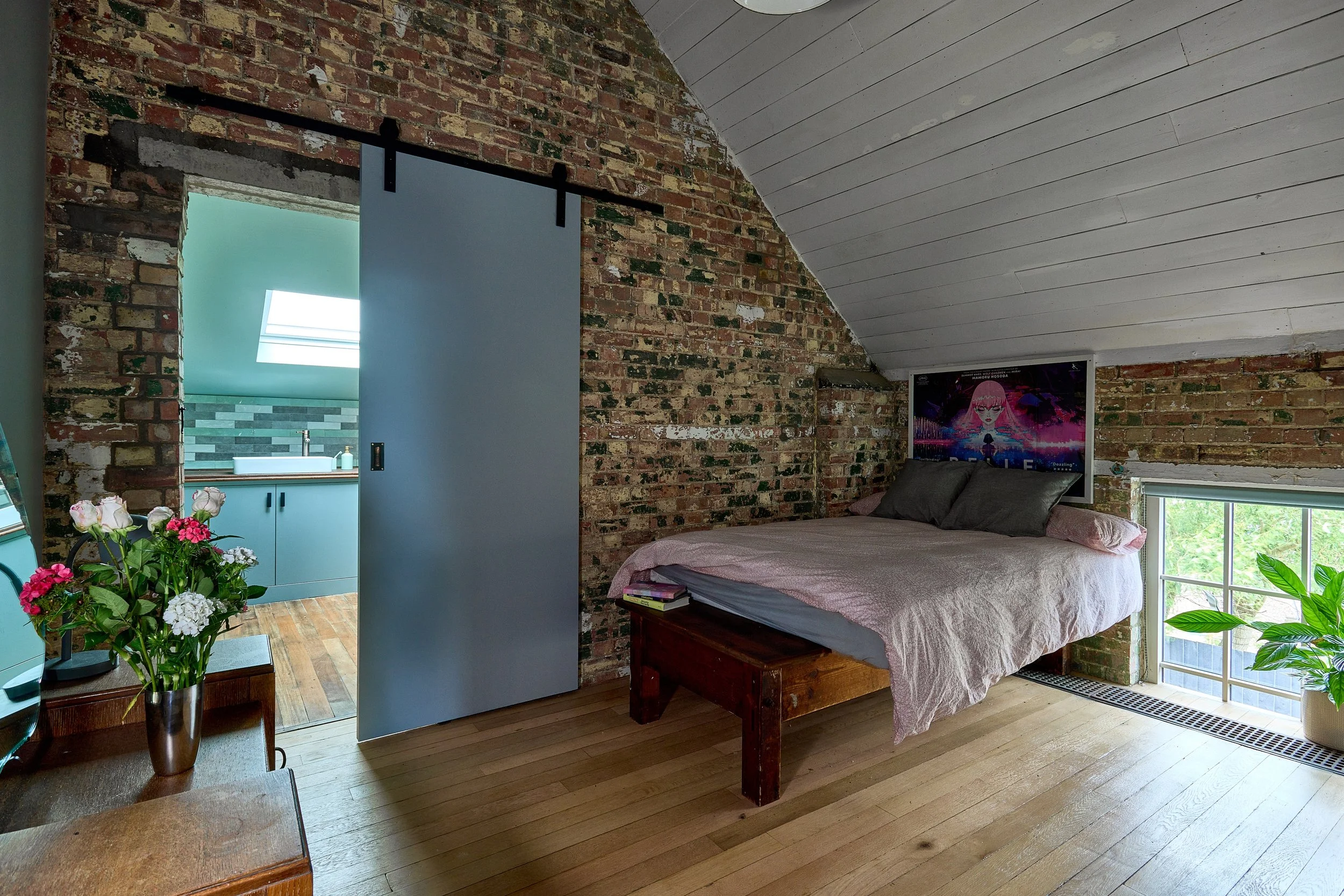 A cozy bedroom with exposed brick walls, a sloped ceiling, a bed with pink and gray bedding, a window with greenery outside, a TV mounted on the brick wall, a wooden bench, and a small foyer leading to a bathroom with a skylight illuminating the spac
