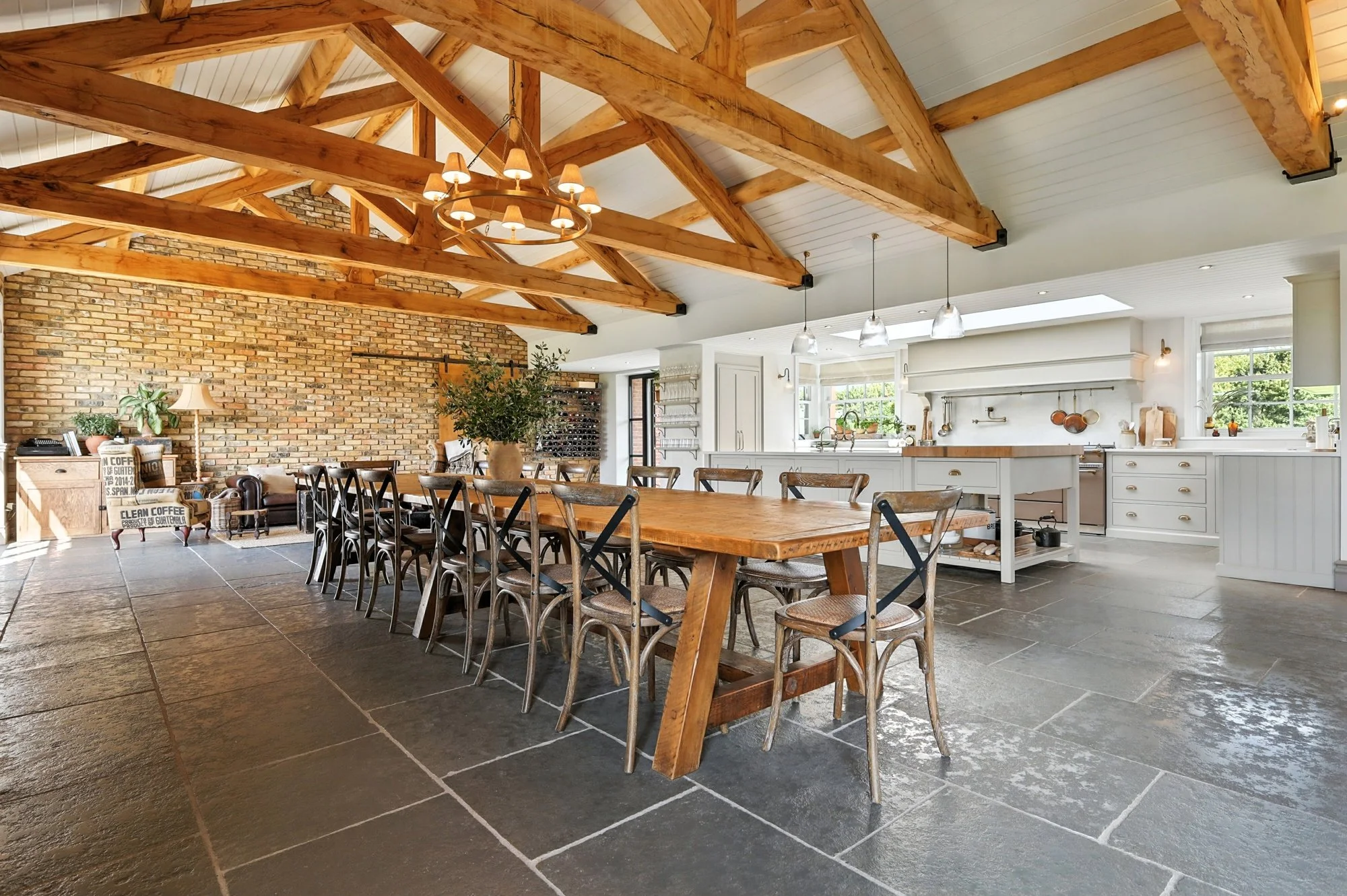 A spacious kitchen and dining area with a high, vaulted wooden ceiling, exposed wooden beams, and a chandelier. The space features a large wooden dining table surrounded by chairs, a brick accent wall, and white cabinetry. There are multiple windows 