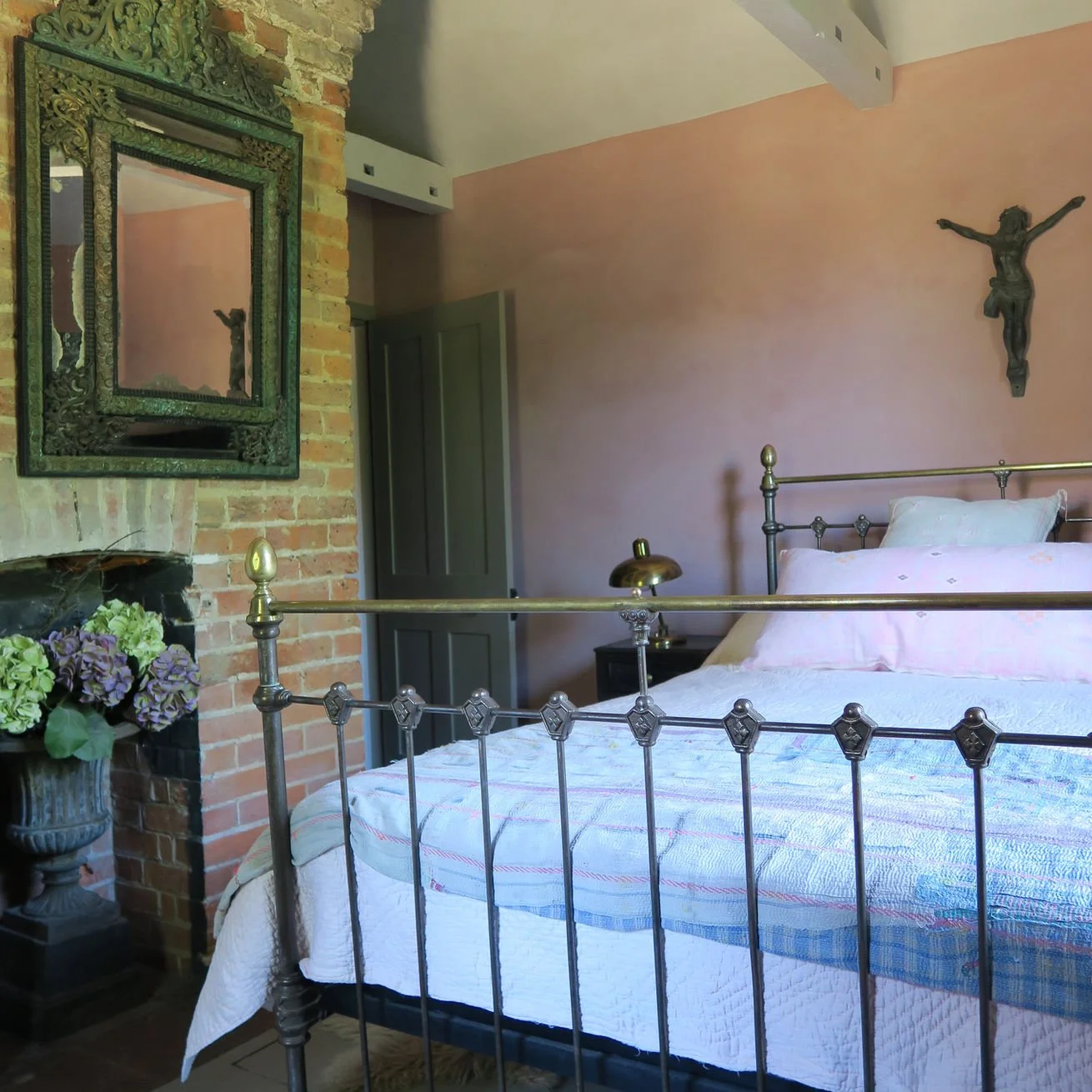 A bedroom with a brass bed, pink bedding, and a quilt. There is a wrought iron crucifix on the pink wall, a lamp on a nightstand, a mirror over a brick fireplace, and a vase of flowers on a side table.