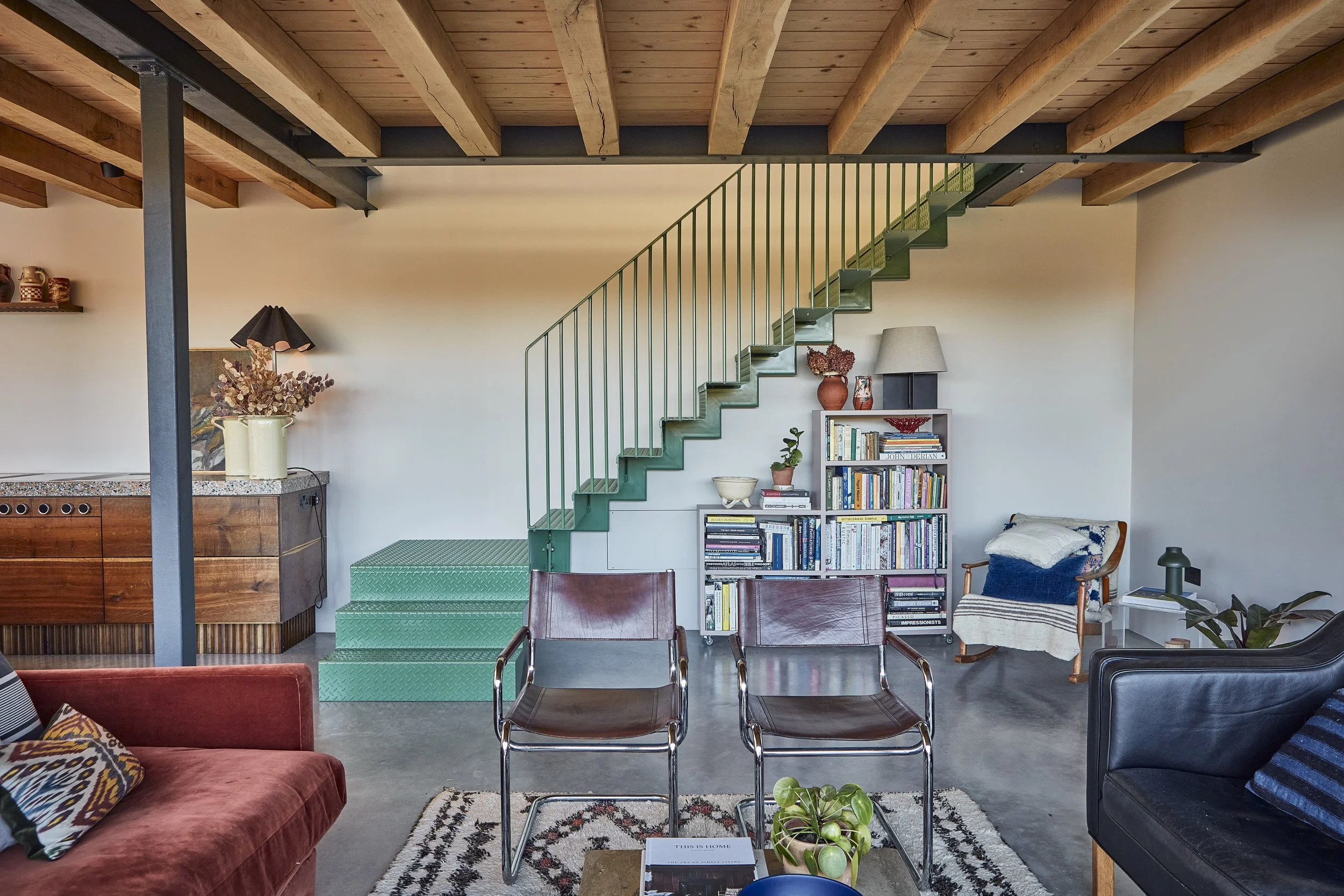 Living room with a wooden ceiling, a green staircase, a bookshelf, various chairs, a red sofa, and plants.