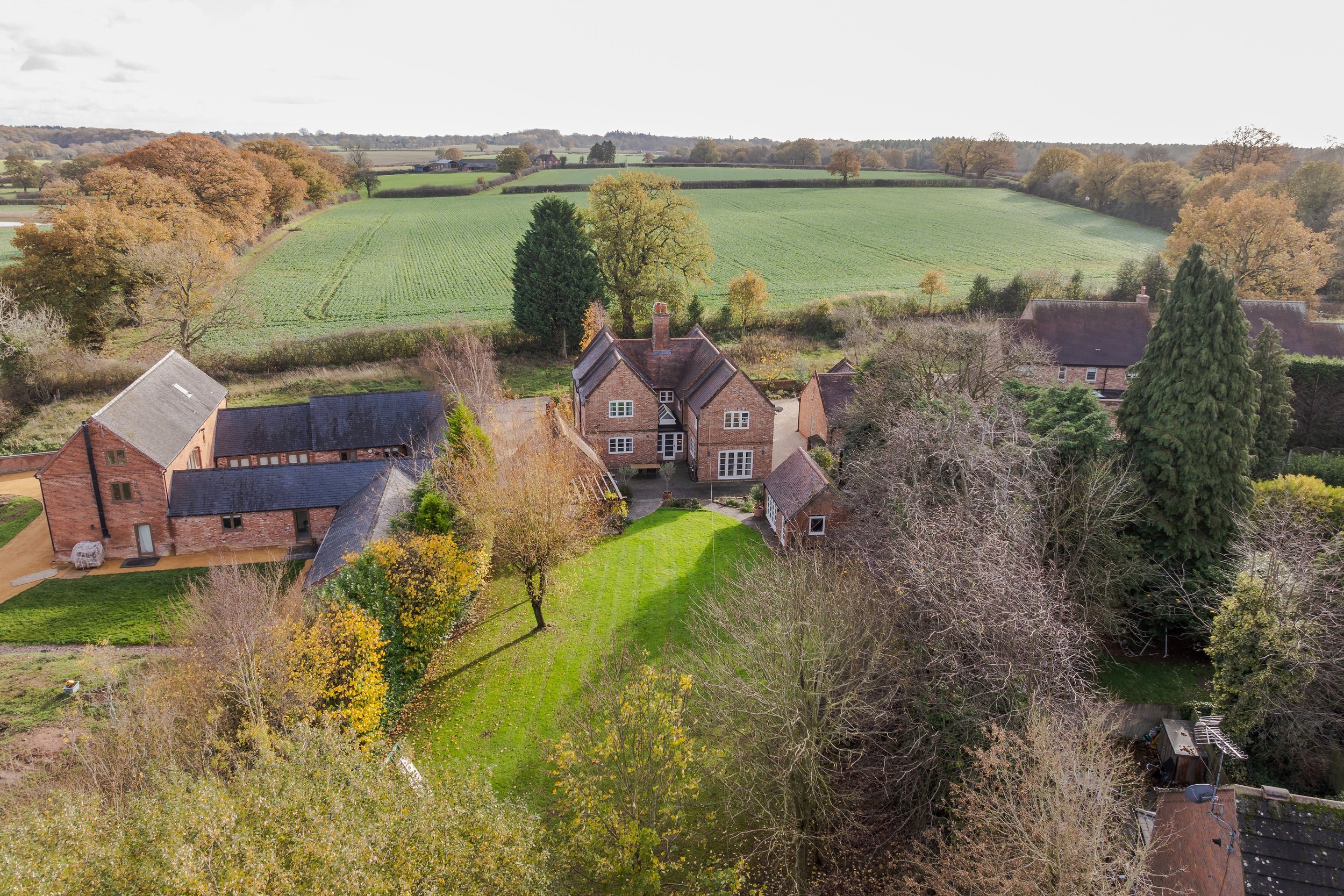 Aerial view of a rural property with a large brick house, surrounded by trees, lawns, and other buildings, with fields and farmland in the background.