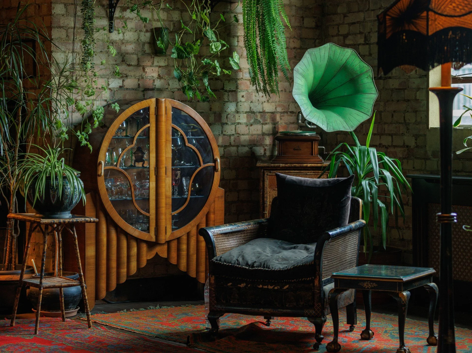 Indoor vintage room with brick wall, surrounded by green plants, wooden furniture including a cane armchair with a pillow, a small table, and a glass display cabinet with glassware. A large vintage gramophone with a green horn is positioned near a wi