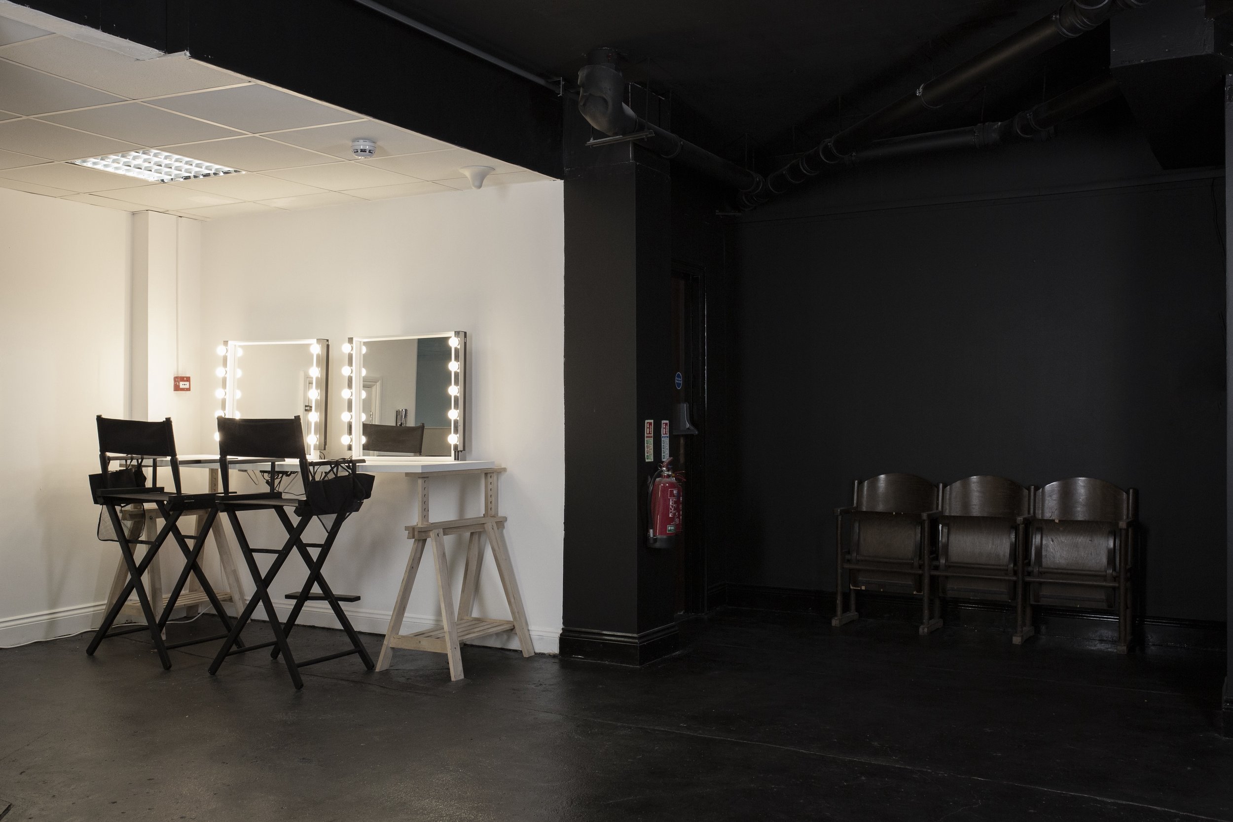Empty backstage area with two illuminated makeup mirrors on the left, and three wooden chairs stacked on the right, with a black wall and fire extinguisher in the background.
