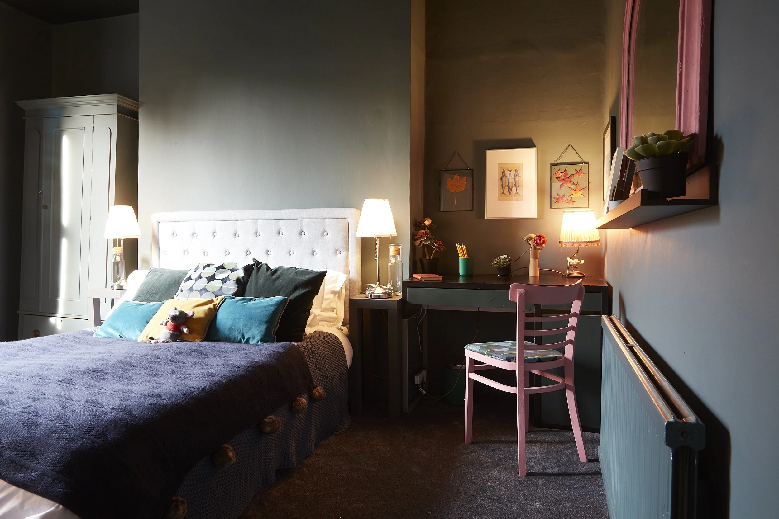 A cozy bedroom with a bed, stuffed animal, pillows, bedside lamps, a desk with colorful decorations, and framed artwork on the wall.