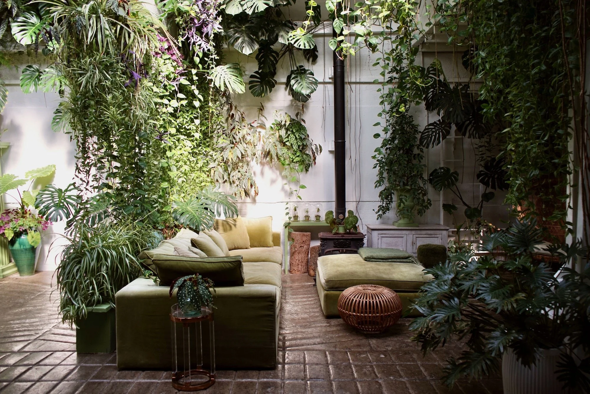 Indoor patio with green sofa, plush pillows, ottoman, wicker stool, surrounded by lush green plants and vines, with a brick floor.
