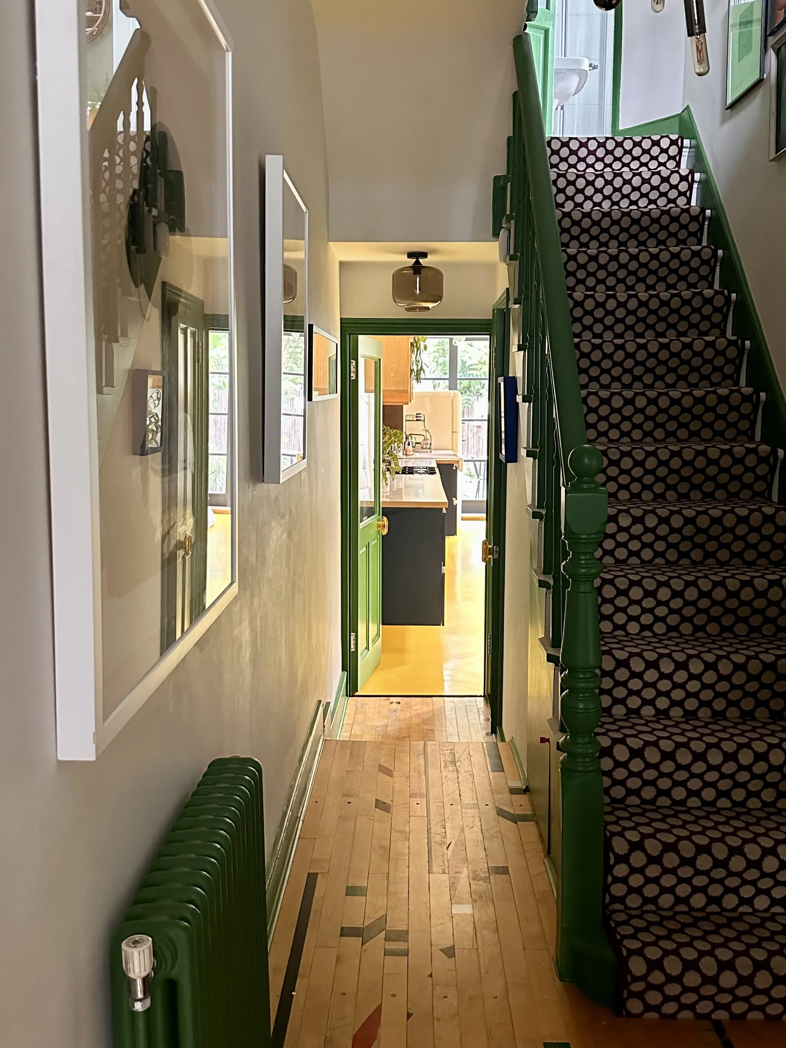 View down a hallway leading to a kitchen with a partly open green door. Stairs with patterned carpet on the right side, framed photographs on the wall on the left, and a green radiator beneath the photographs.