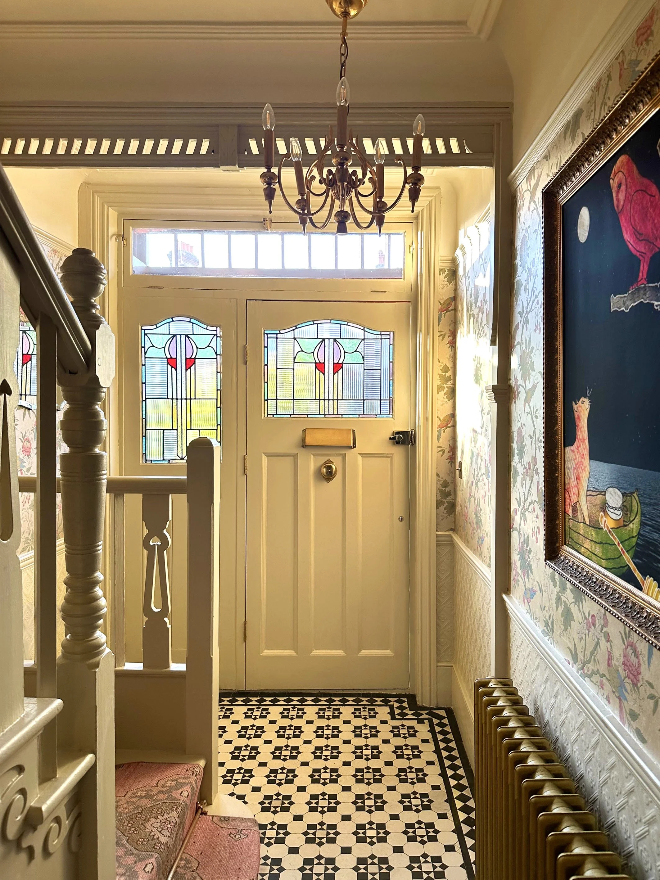 Entryway with vintage decor, including a stained glass front door, floral wallpaper, chandelier, a small pink patterned rug, and a radiator.