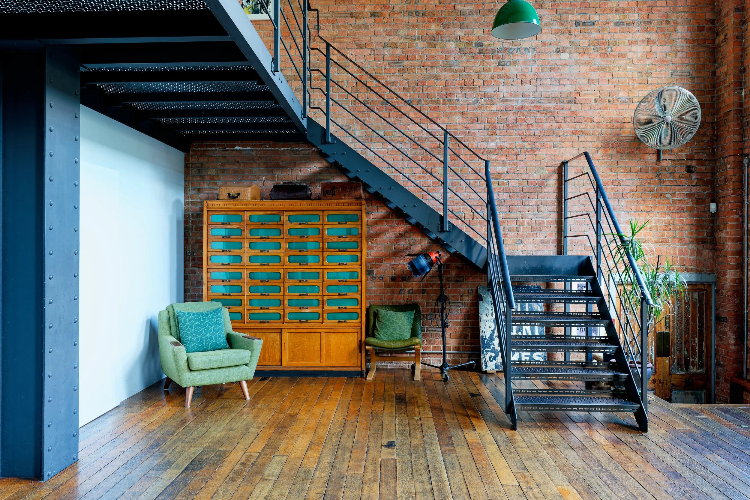 Interior of loft with brick walls, wooden floor, black metal staircase, green armchair, wooden cabinet, a small chair, and wall fan.
