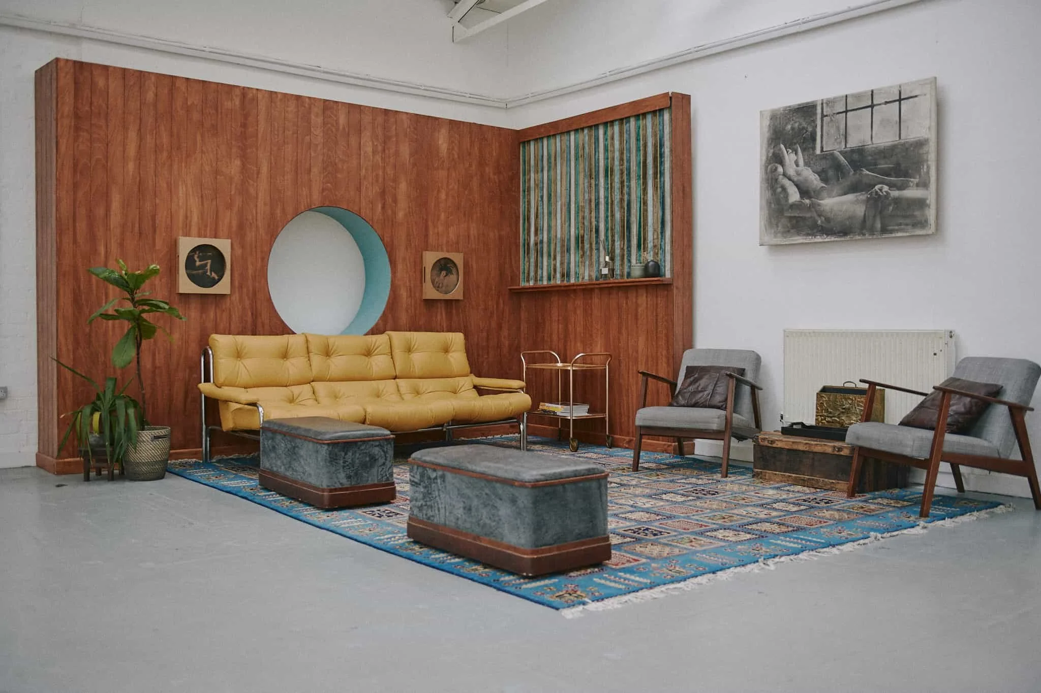 A stylish living room with a wood-paneled accent wall, a yellow tufted sofa, two gray ottomans, and two gray armchairs. There is a large framed black-and-white painting on the white wall, a potted plant in a basket, and a colorful rug on the light gr