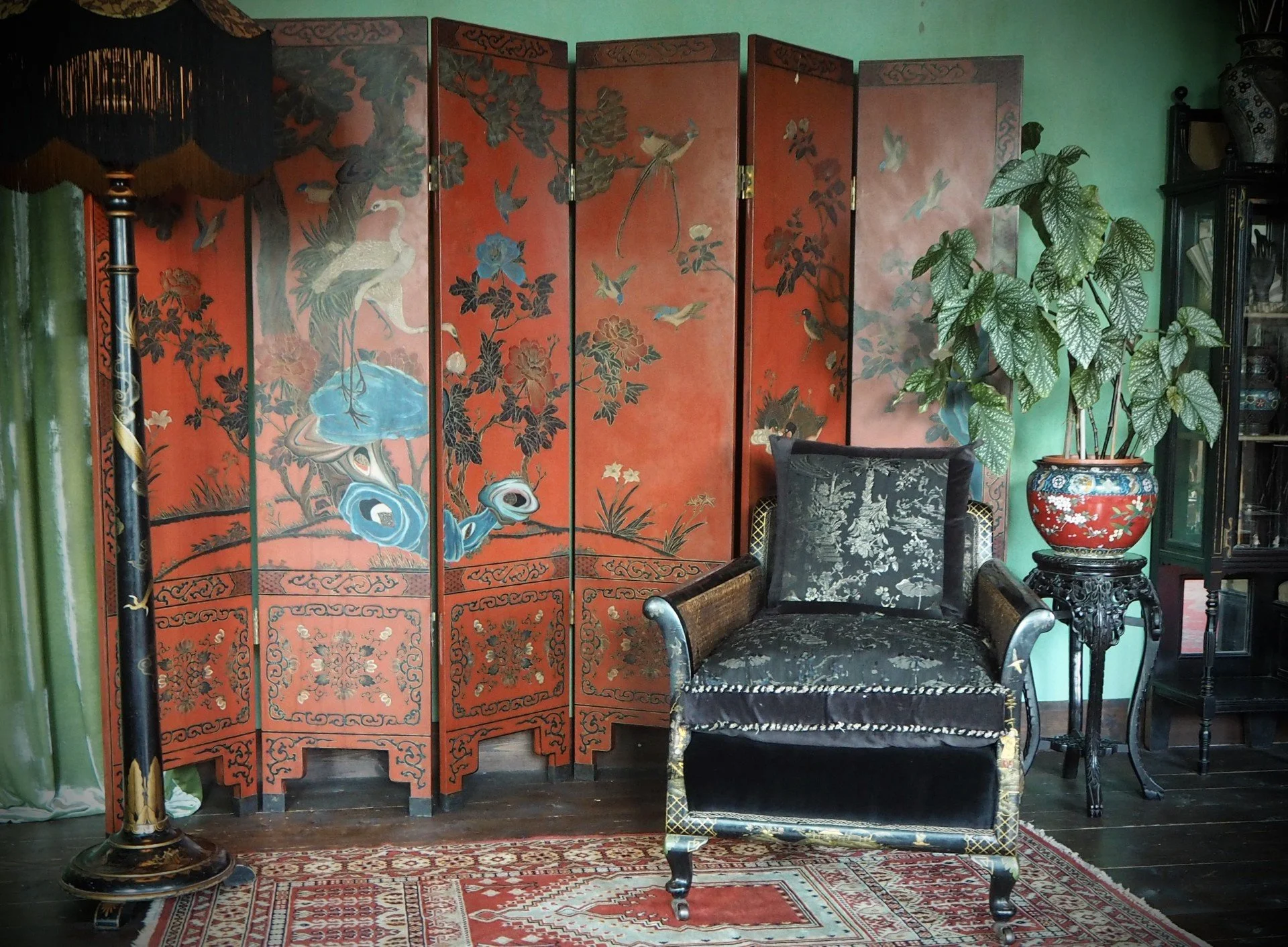 An ornate black velvet armchair with a floral pattern, positioned in front of a decorative red folding screen with Chinese-style artwork of birds, flowers, and trees. To the right, there is a large potted plant with variegated green leaves on a black