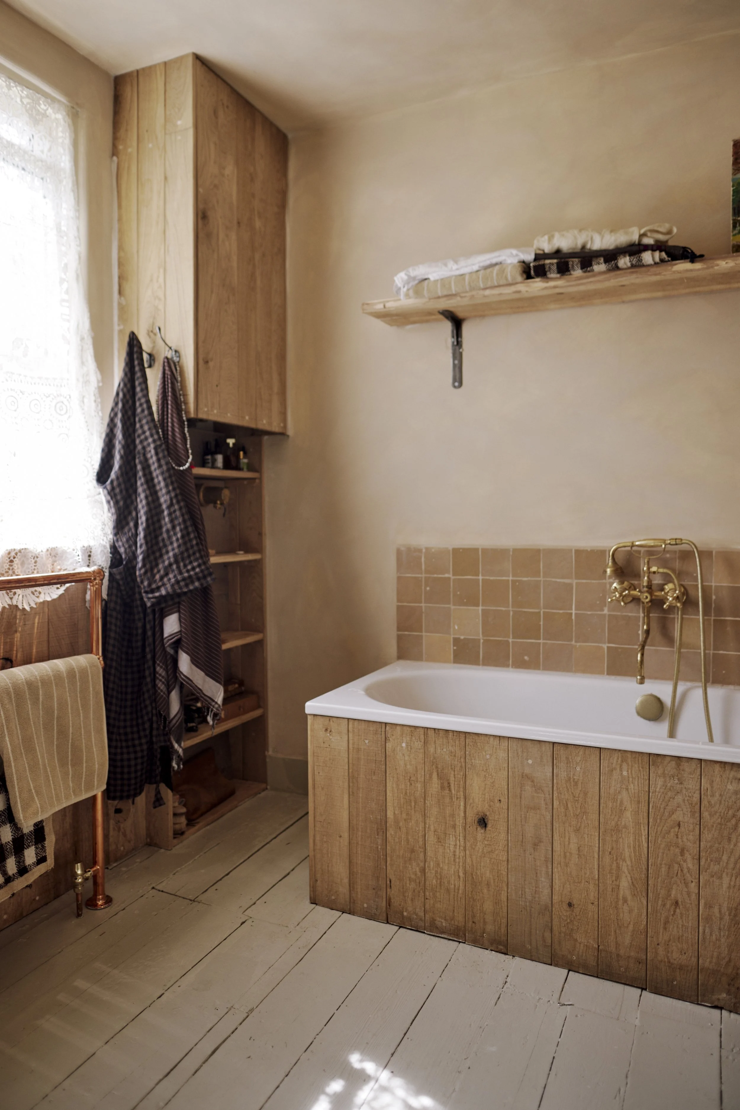 A rustic bathroom with a wooden bathtub, a brass faucet, a wooden shelf with folded towels, a wooden cabinet with hanging checkered aprons, a window with lace curtains, and a towel rack with a plaid towel.