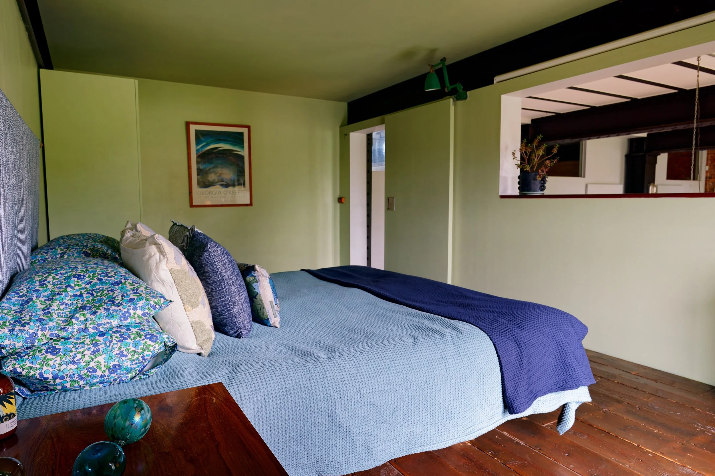 A bedroom with a bed covered in a blue quilt, multiple pillows, and a wooden nightstand with decorative glass objects.