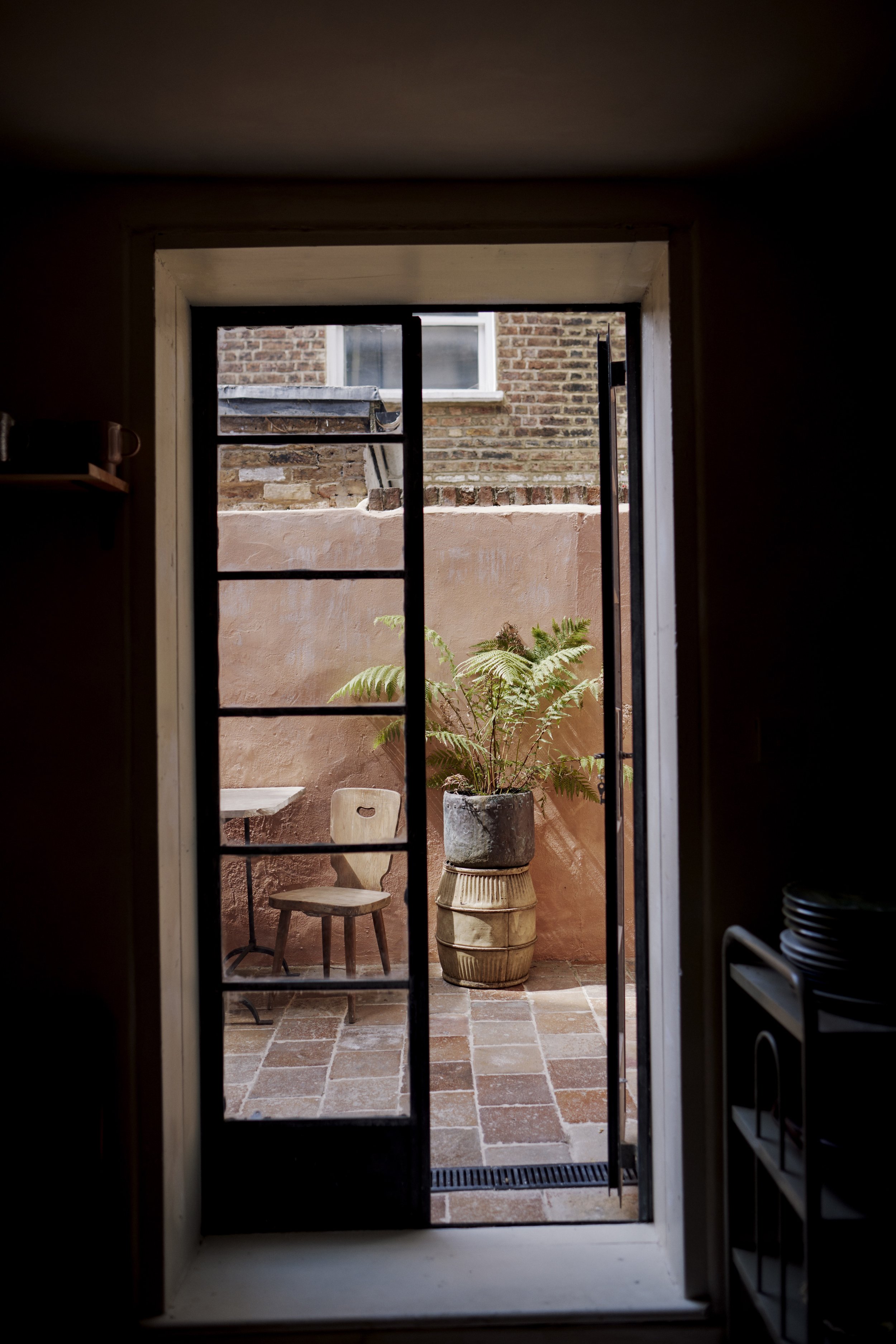 View through an open glass door revealing a small outdoor patio with a potted fern, an old wooden chair, and a brick and stucco wall, with neighboring brick building in the background.