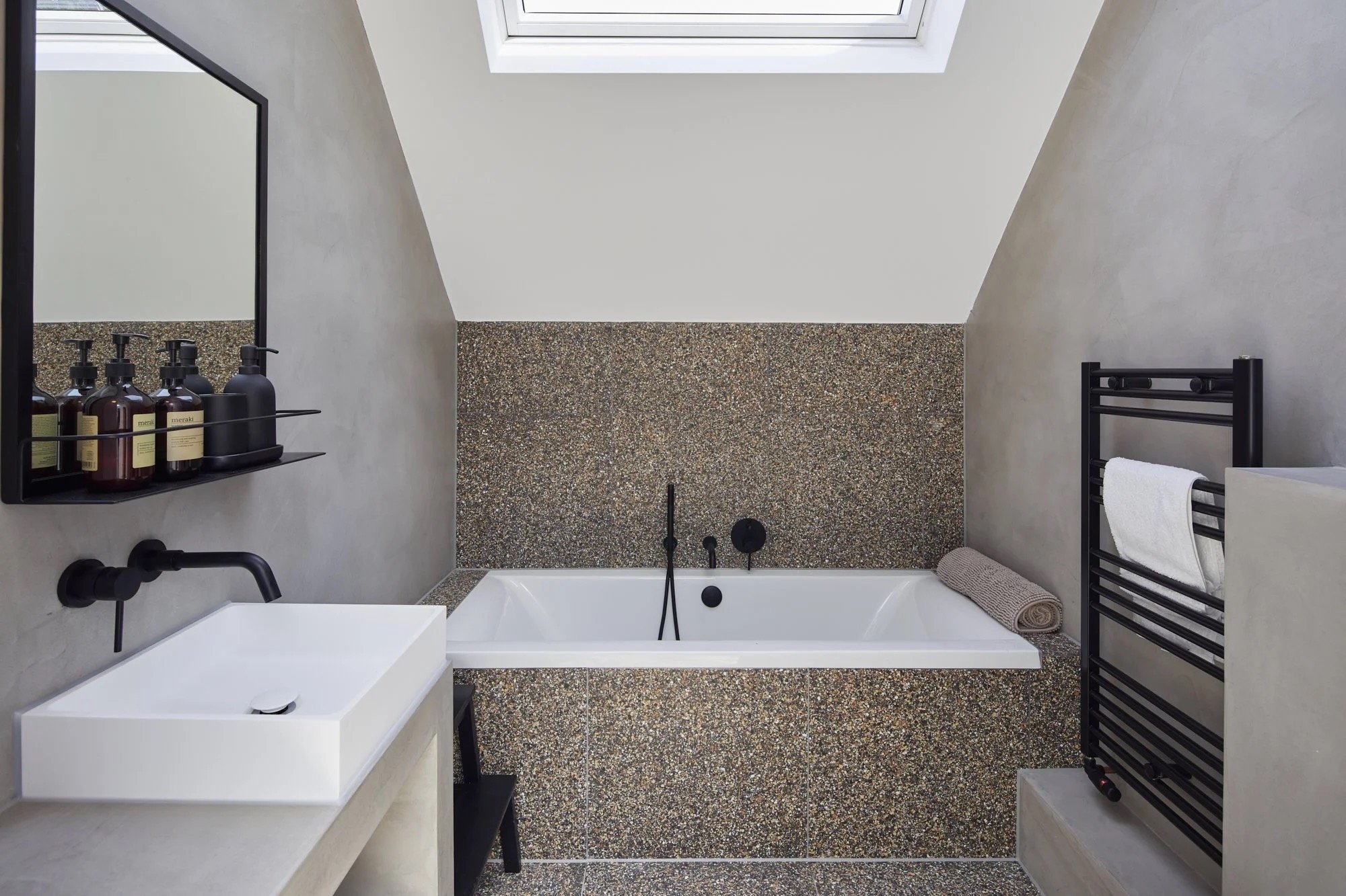 Modern bathroom with a bathtub under a skylight, black towel rack with a white towel, floating vanity with a black faucet, and a black shelf with toiletries.