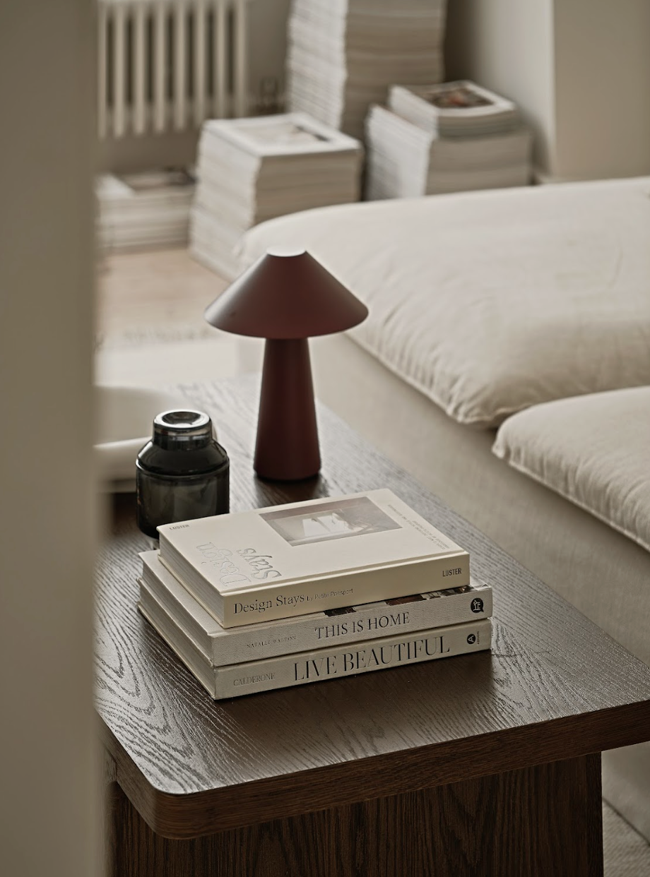 A wooden bedside table with a stack of four books, a dark-colored jar, and a table lamp with a conical shade in a cozy living space or bedroom.