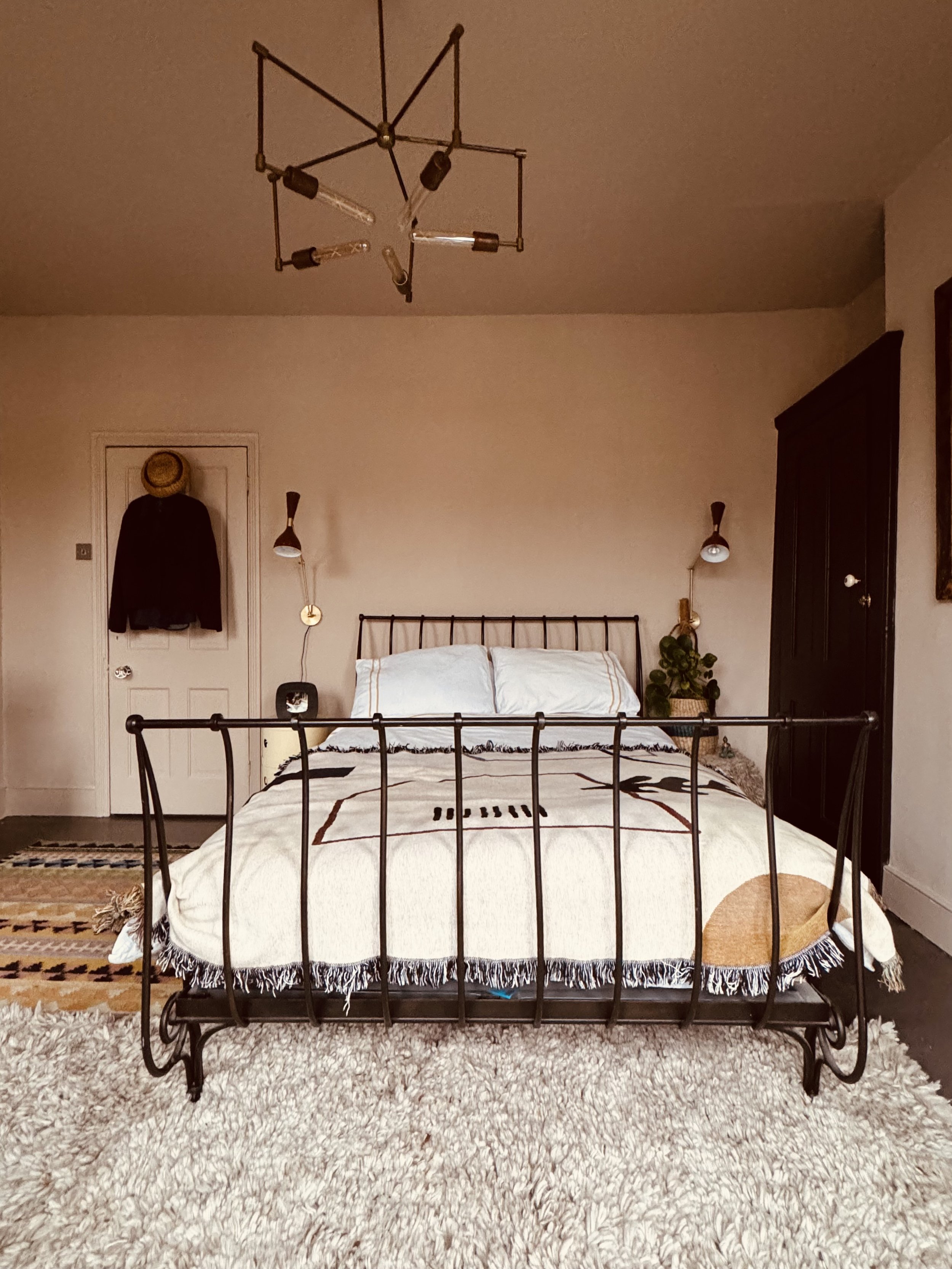 A bedroom with a bed featuring a black metal frame, a white bedding, and two pillows. There are wall-mounted lamps on either side of the bed, a wardrobe on the right, and a door with a coat and a hat hanging on it. The floor has a beige shaggy rug, a