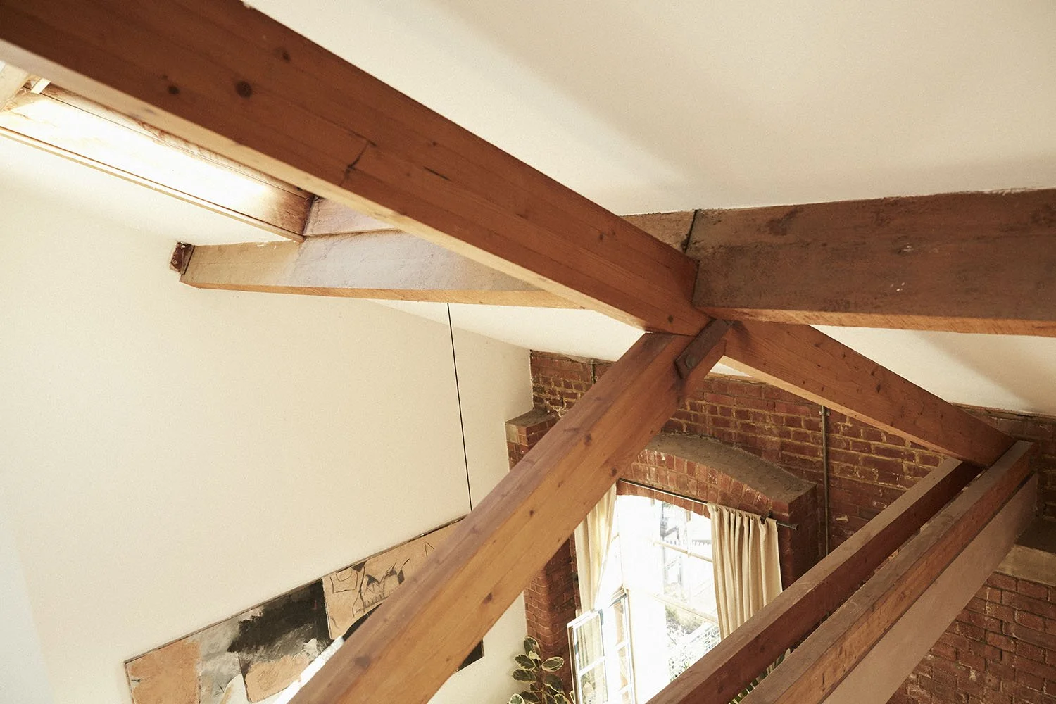 Interior of a room showing wooden beams and brick walls with a window and curtains.