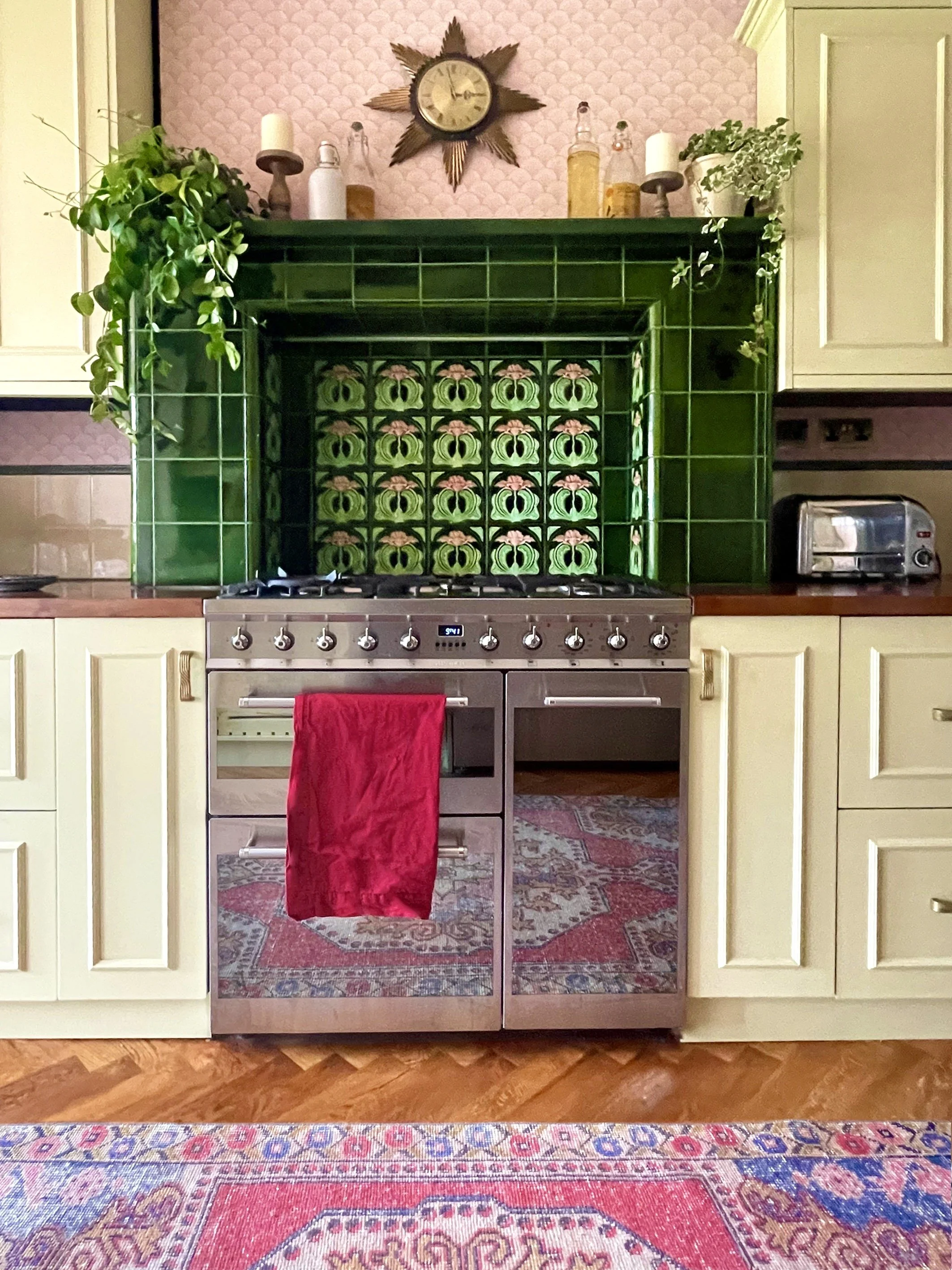 A kitchen with a green tiled fireplace, vintage patterned tiles, and green cabinetry. The fireplace has plants and decorative bottles on the mantle, a star-shaped clock, and a red towel hanging on the oven handle. There's a toaster on the right side 