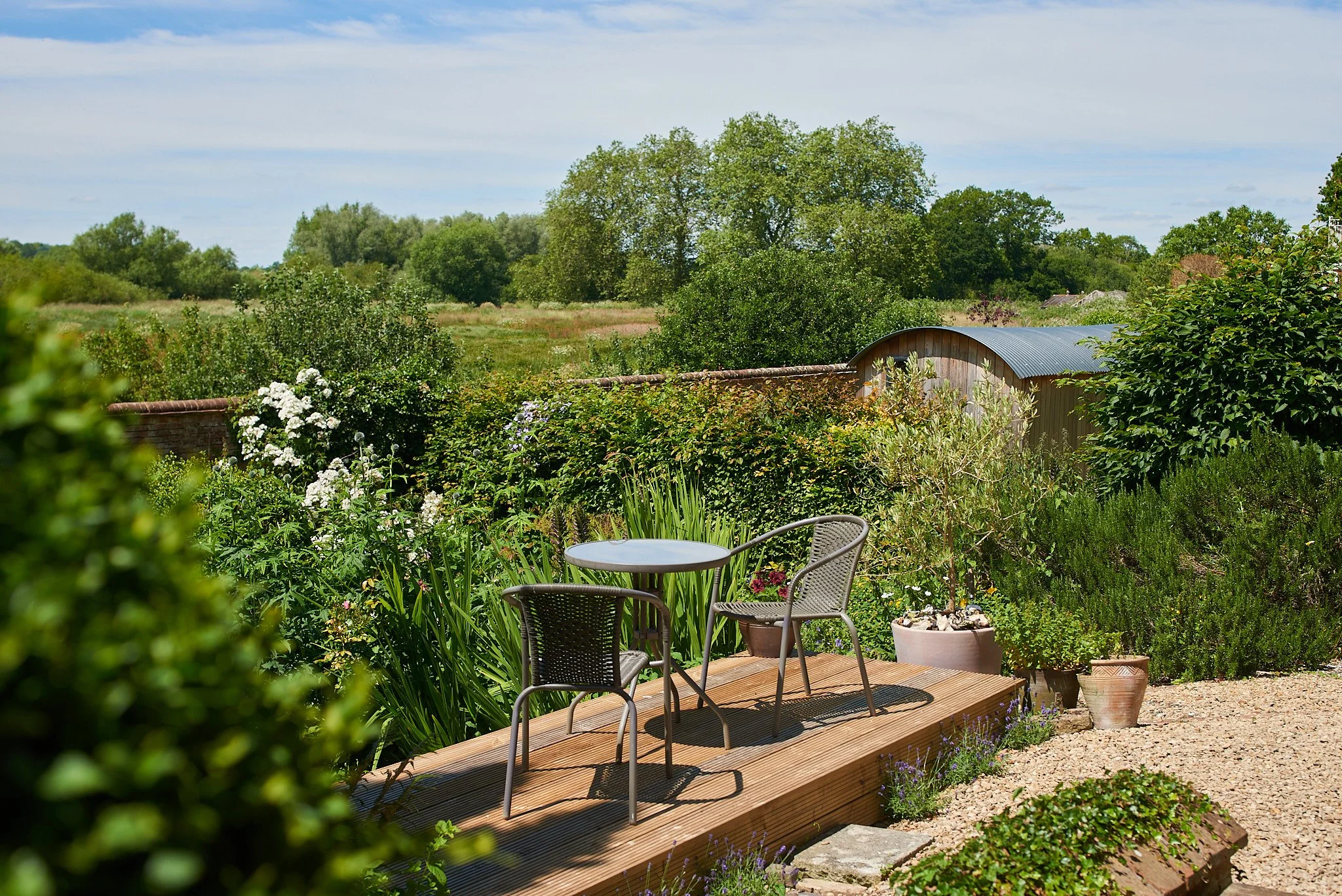 A small outdoor patio with a round table and two chairs on a wooden deck, surrounded by lush green plants and flowers, with a rural landscape and trees in the background under a clear blue sky.