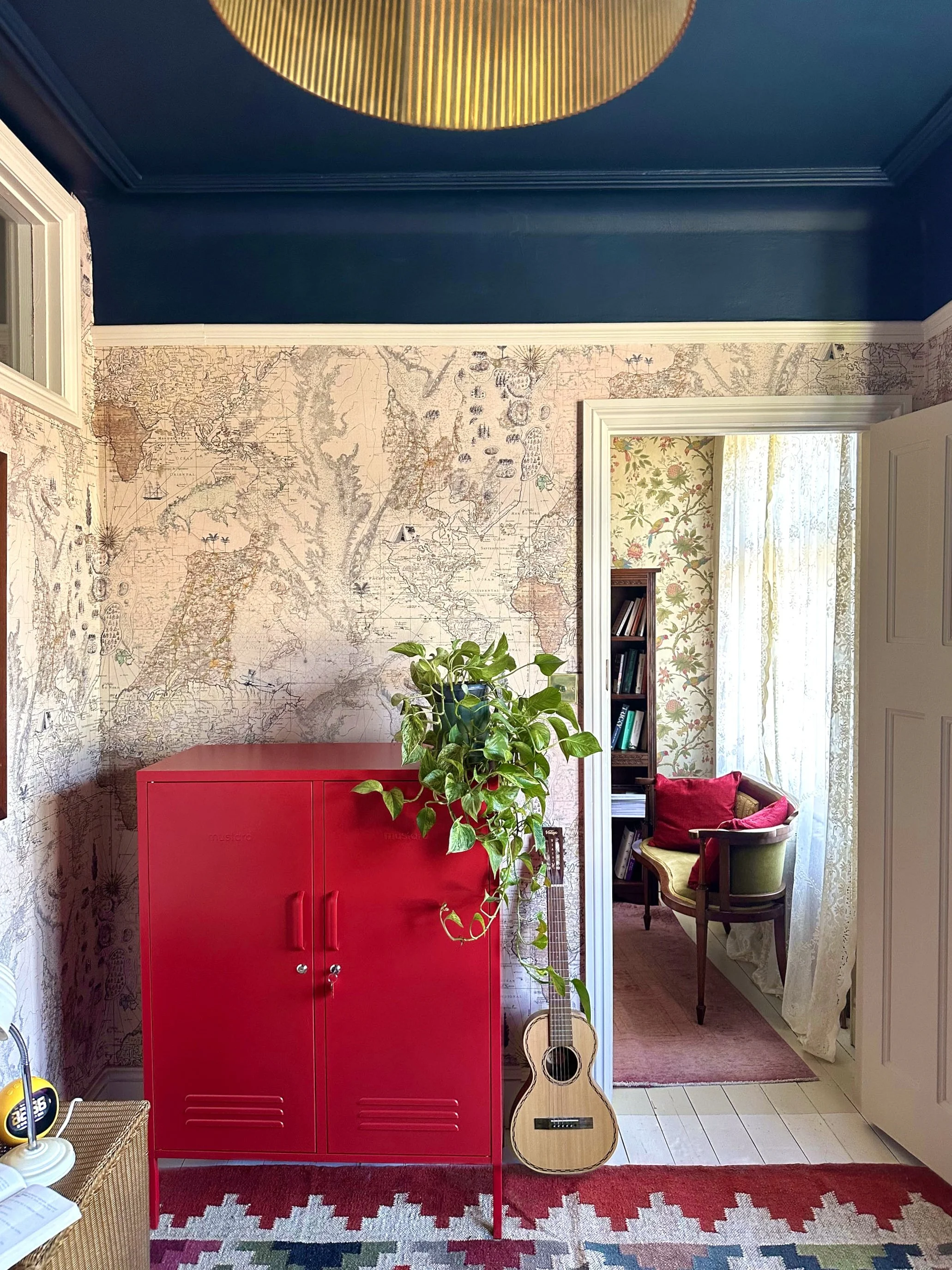 Interior room with a red cabinet, a potted plant, an acoustic guitar, and a large wall map. A doorway reveals a cozy sitting nook with a window, lace curtains, and a bookshelf.