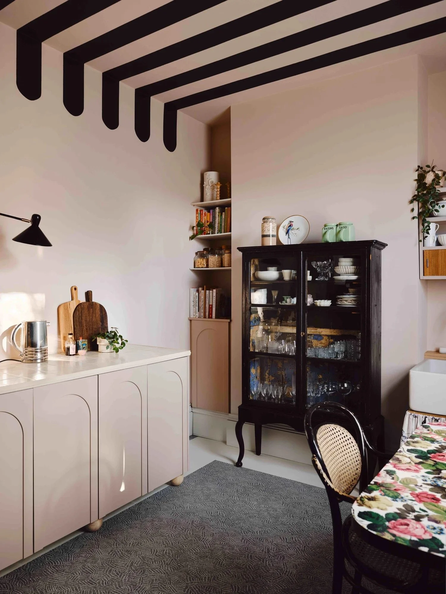 A dining area with a floral tablecloth and black chairs, a black display cabinet filled with glassware and dishes, a white sideboard, and a small bookshelf with decorative items and plants. The ceiling has black and beige striped beams.
