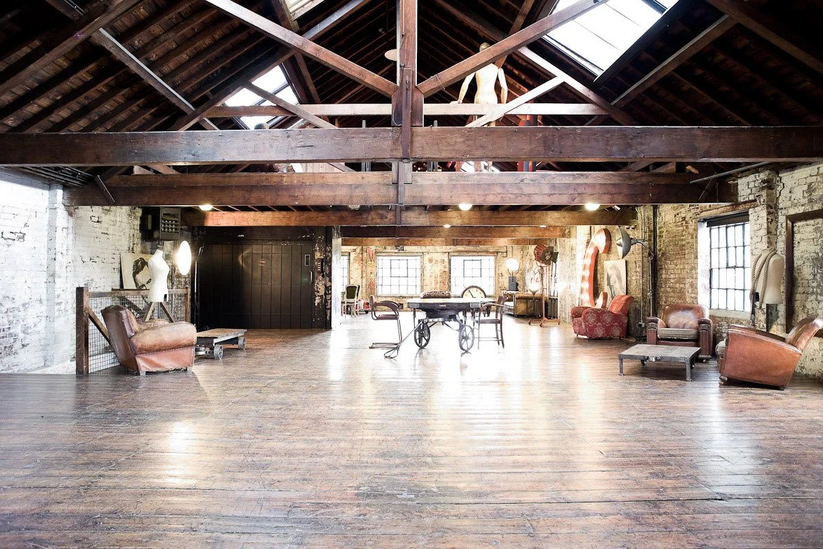 A spacious, rustic loft with exposed brick walls, wooden beams, and hardwood floors. Vintage furniture including armchairs and coffee tables, with large windows allowing natural light to brighten the room.