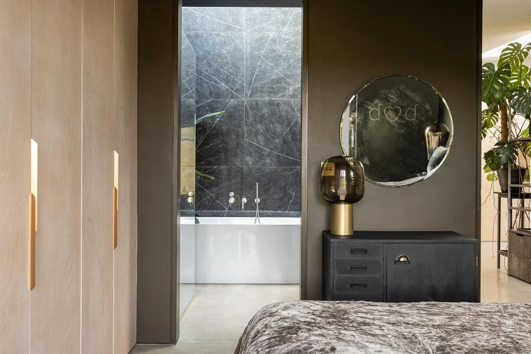 Modern bedroom with a black dresser, gold and black vase, round mirror, and view of a bathroom with black marble walls and a white bathtub.