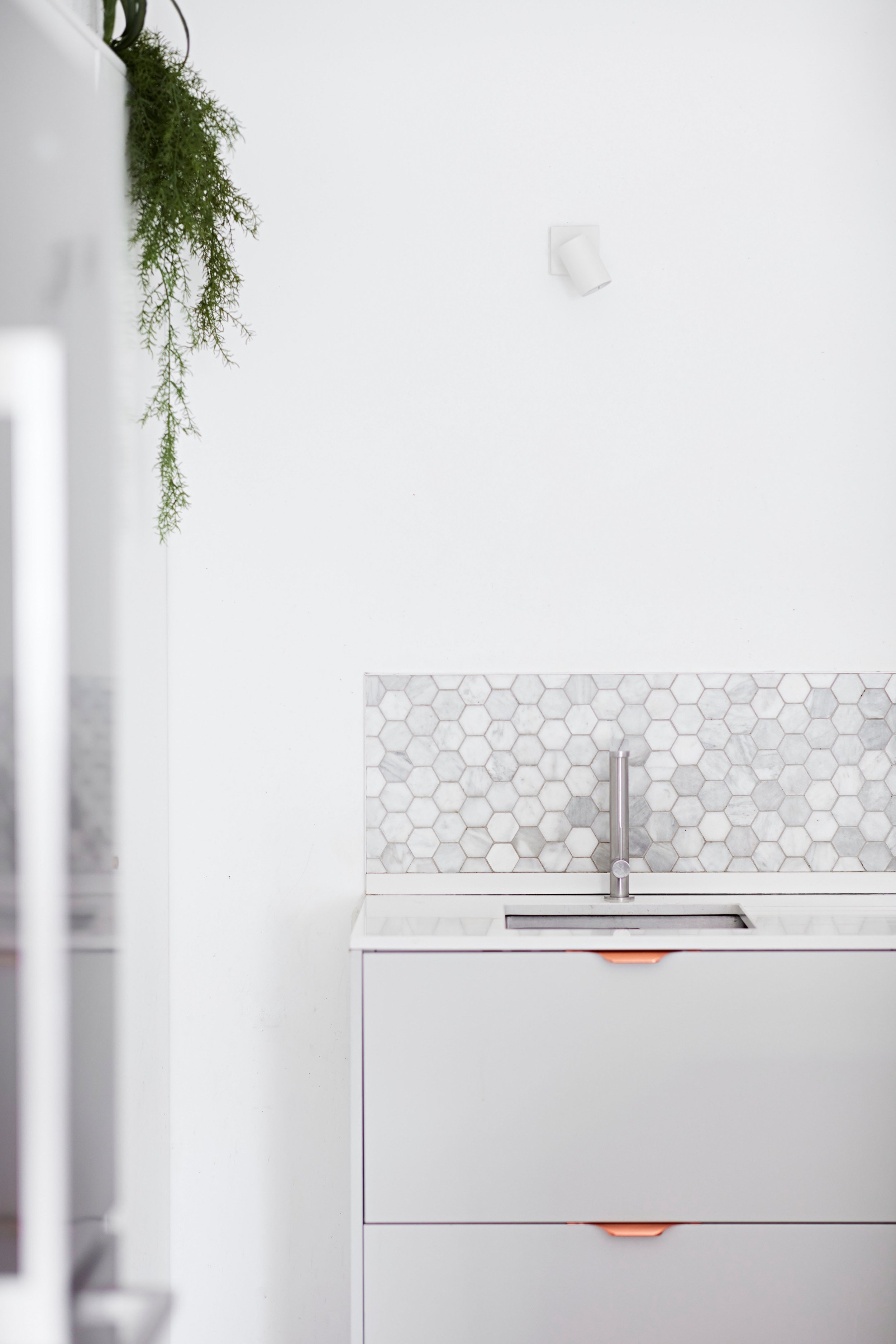 A white kitchen with a mosaic tile backsplash, a silver faucet, and a white cabinet with copper handles. A green hanging plant is visible on the left side.