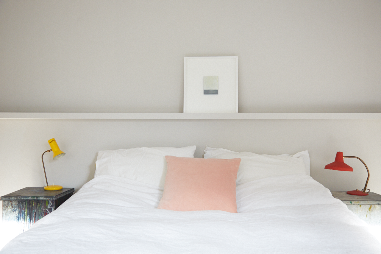 A neatly made bed with white bedding, a pink pillow in the center, and two small bedside tables with colorful lamps, one yellow and one red. There is a framed abstract artwork on the wall behind the bed.
