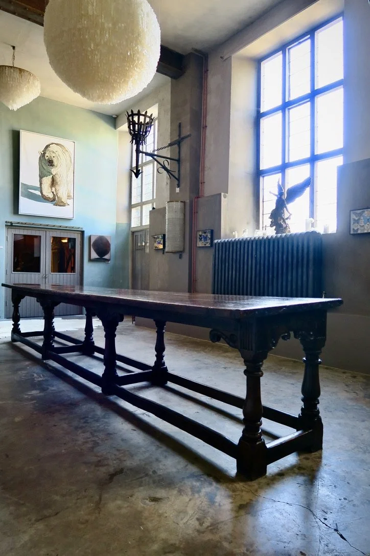 An industrial-style interior room with large windows, a long wooden table, hanging light fixtures, and various decorative art pieces on the walls, including a painting of a bear.