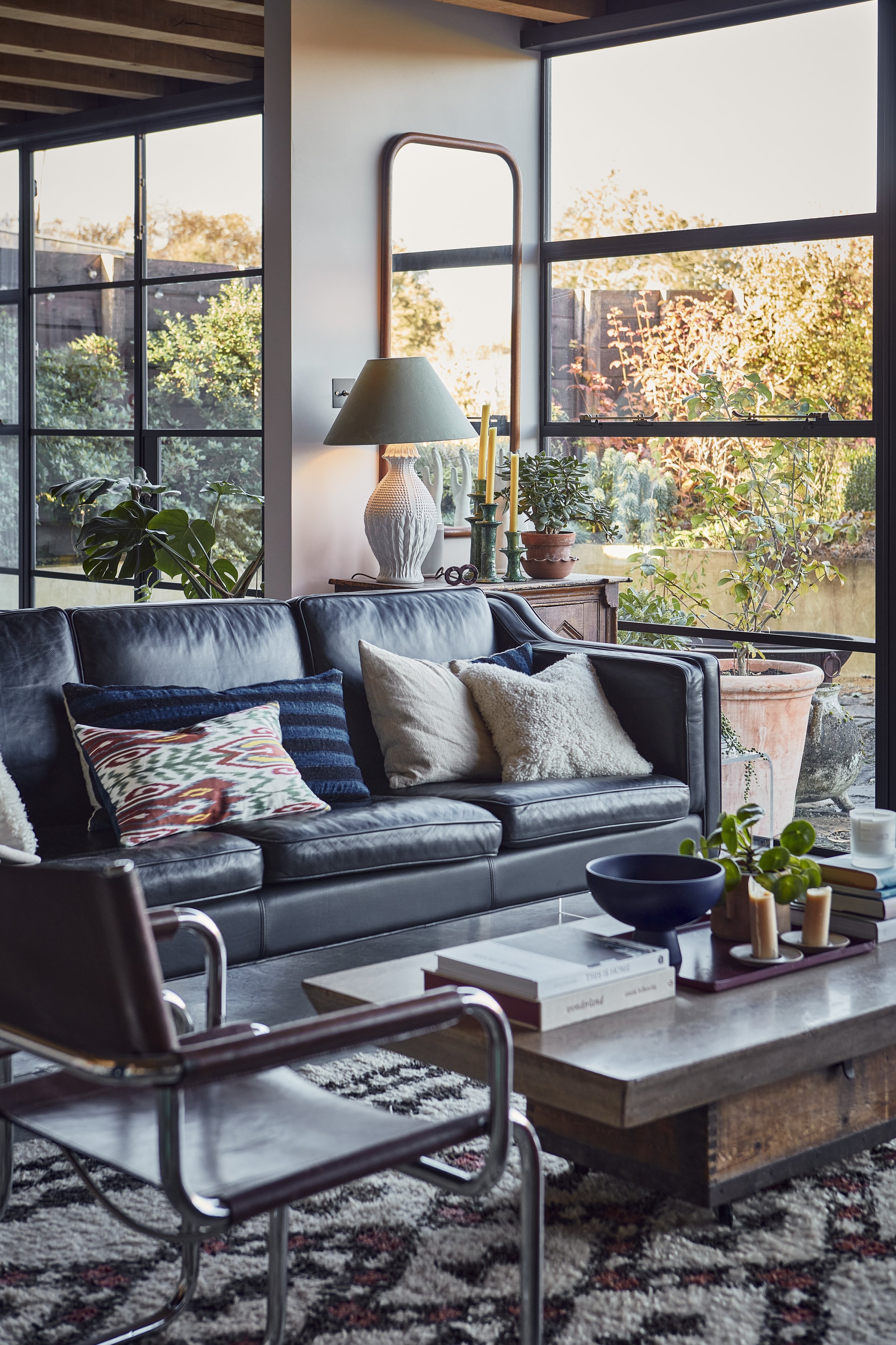 Living room with a black leather sofa, colorful pillows, a wooden coffee table with books and candles, a large window with a view of trees, and decorative plants and lamps.