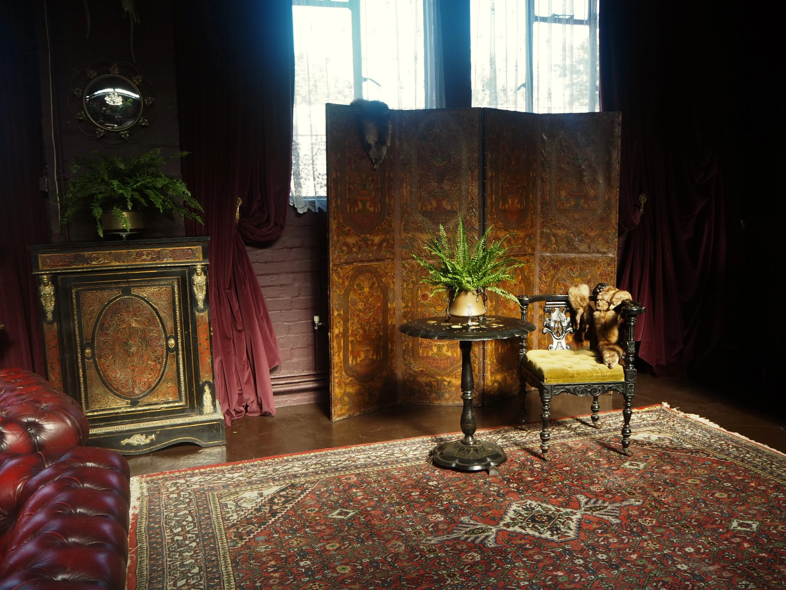 Vintage interior room with red leather sofa, ornate cabinet with gold accents, folding decorative screen, potted fern, and an antique chair with a faux fur blanket, rug, and curtains.