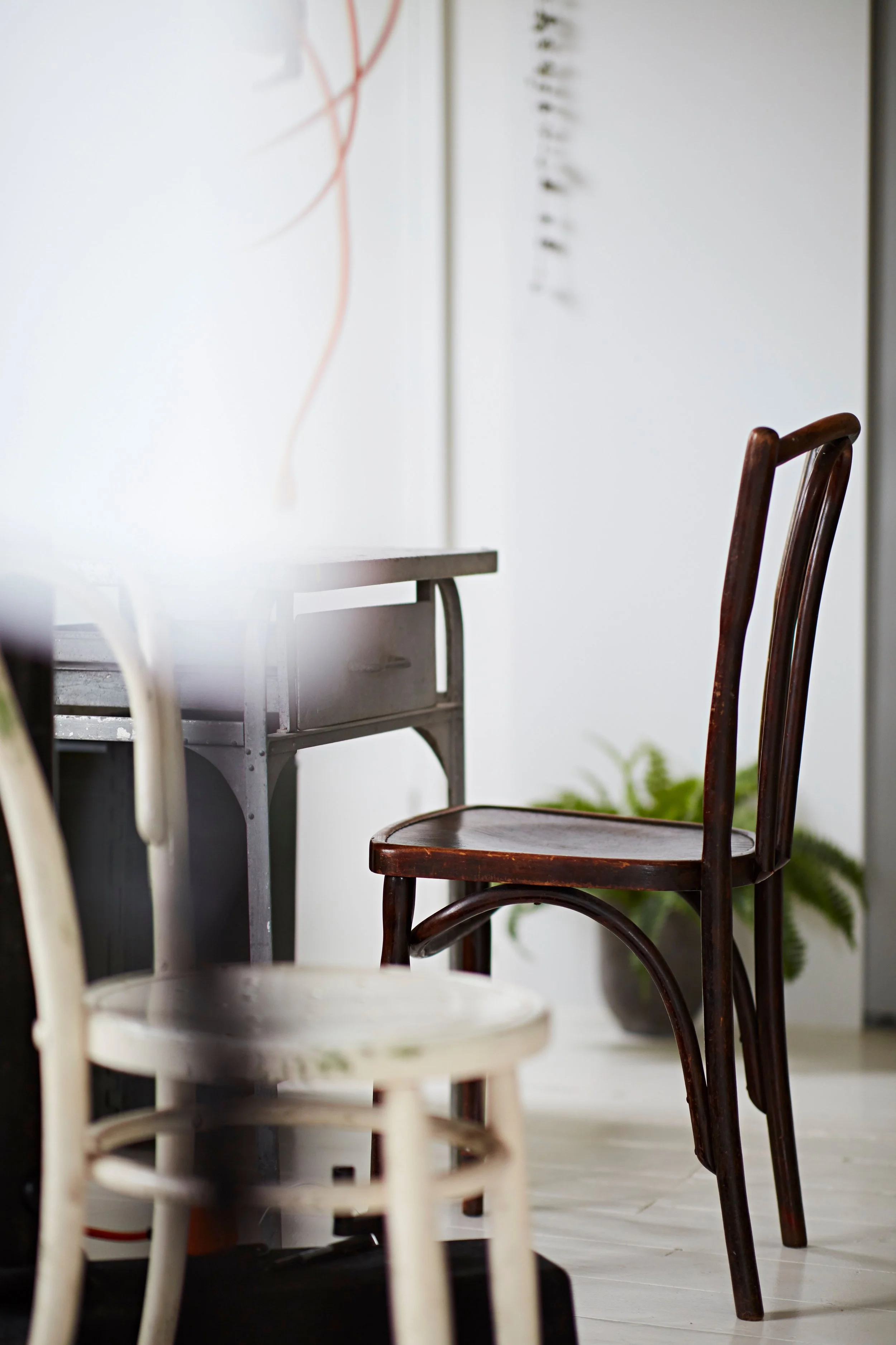 Interior room with a white table, brown wooden chairs, and a white chair with a worn finish. A potted plant is on the floor near the wall, and part of a white wall with some abstract artwork is visible.