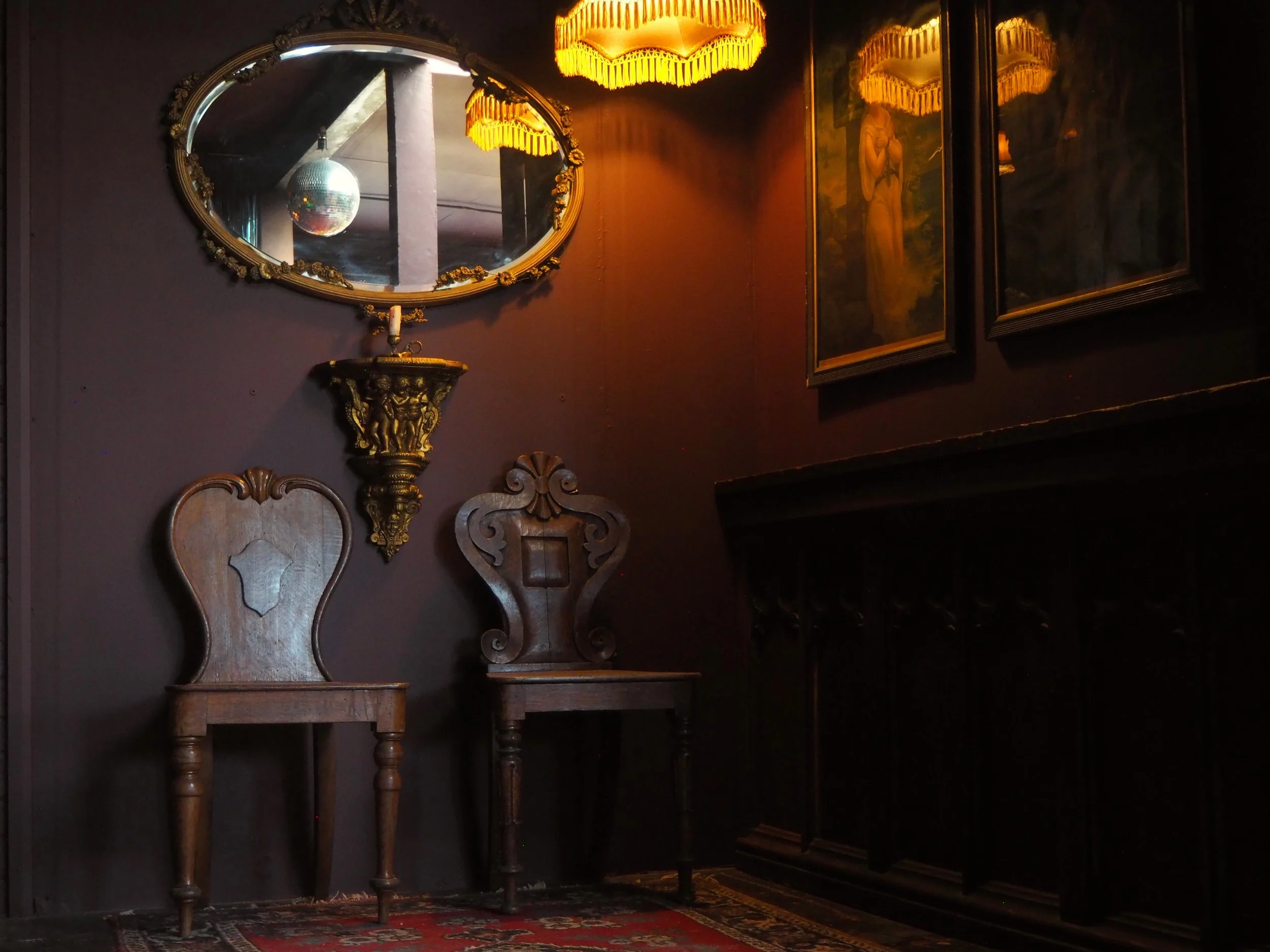 A dimly lit vintage room with a dark wooden sideboard, two ornate wooden chairs, a mirror with an oval frame, and framed paintings. The mirror reflects a partial view of a ceiling with a disco ball. Warm yellow lighting comes from two fringed lampsha
