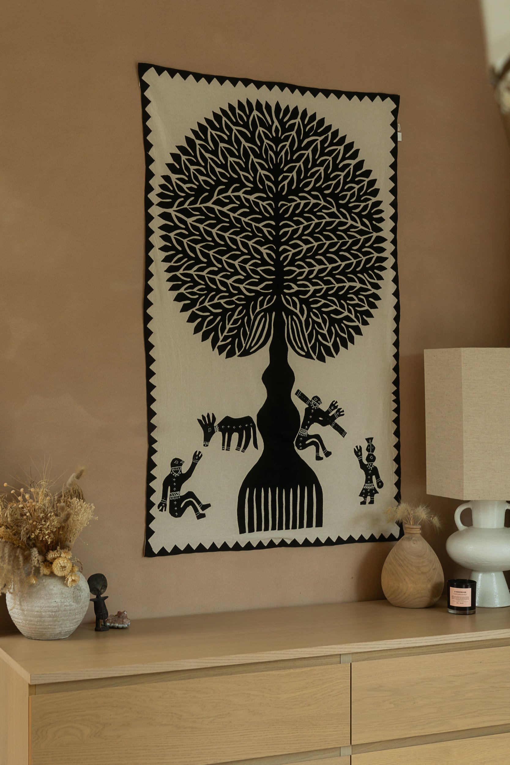 Wall hanging artwork with a black tree and figures on a beige background on a wooden dresser.