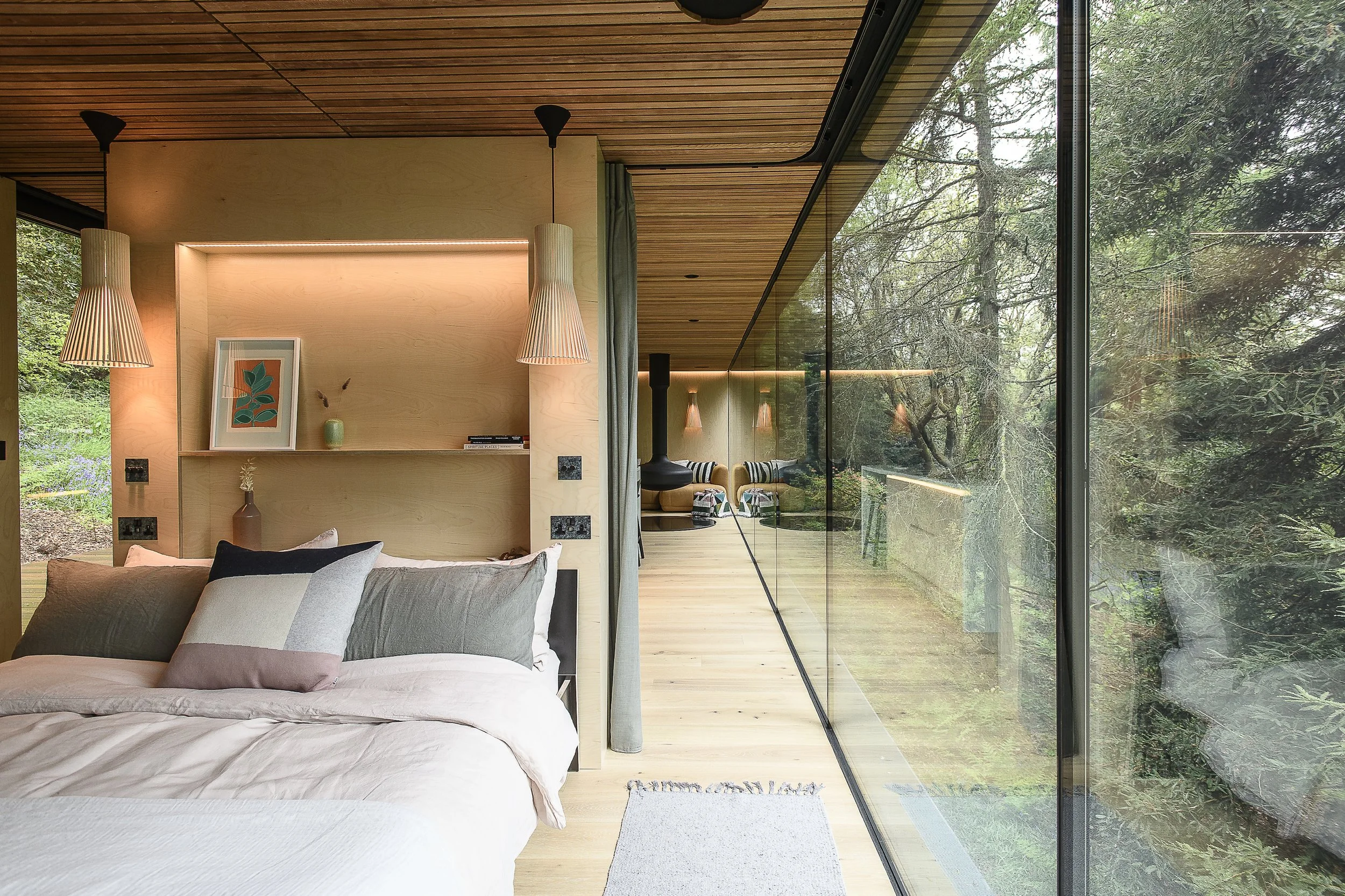 Modern bedroom with a large glass wall overlooking a forest, featuring wooden ceiling and floors, a bed with cushions, hanging lamps, and decorative items on shelves.