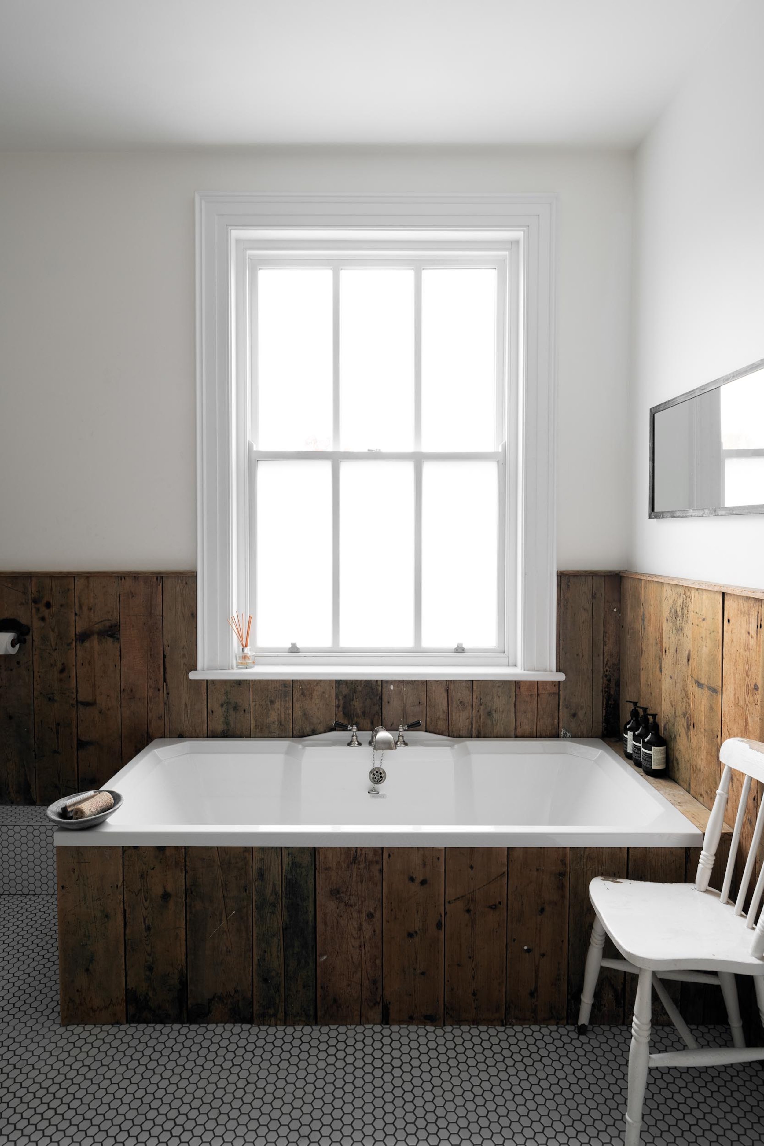 A bathroom with a white bathtub surrounded by wood paneling, a window above the tub, and a white chair beside it.