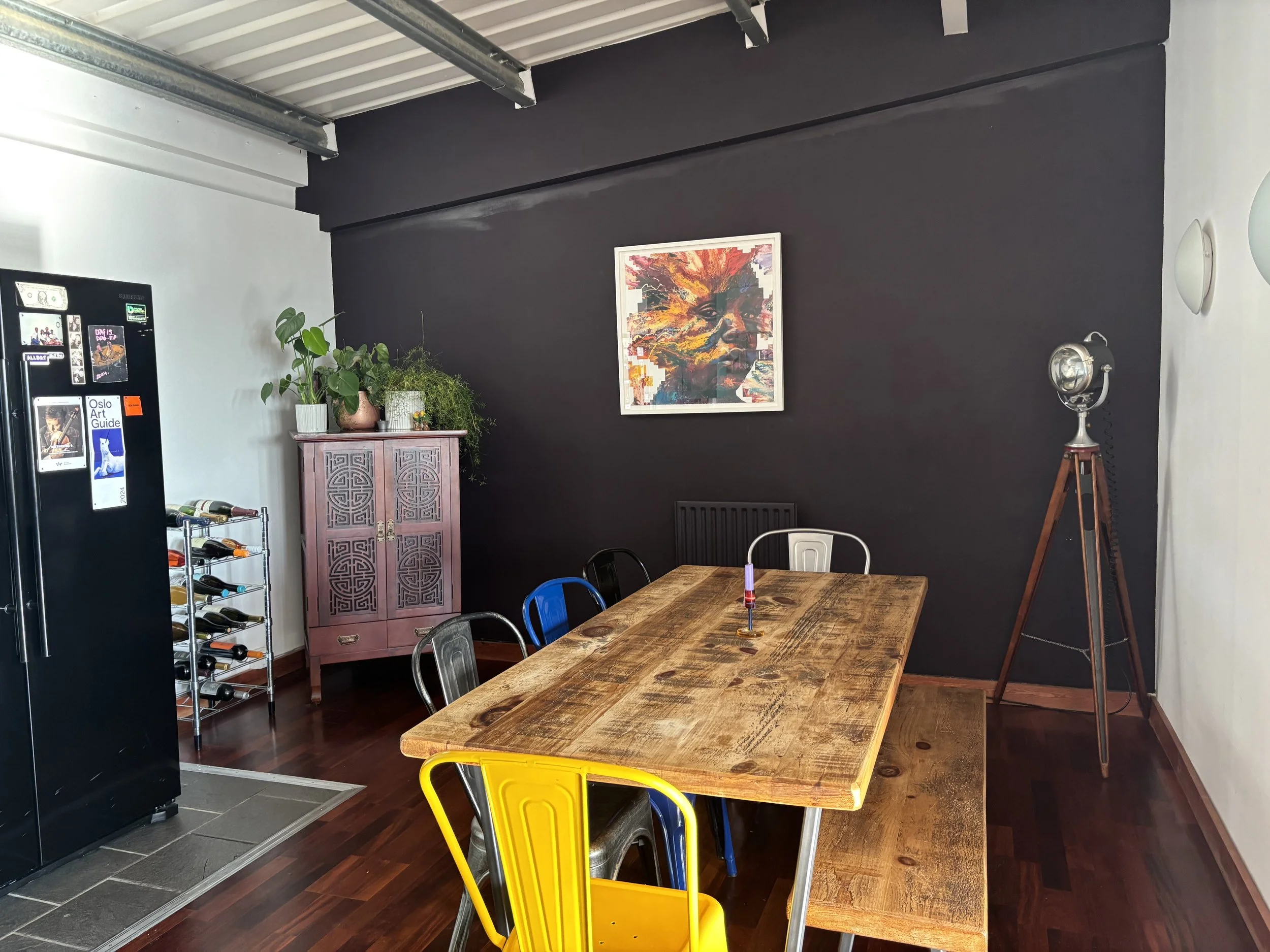 A dining area with a large wooden table, multicolored metal chairs, a black wall with a colorful abstract face painting, a pink cabinet with plants, a wine cooler with wine bottles, and a vintage-style floor lamp.