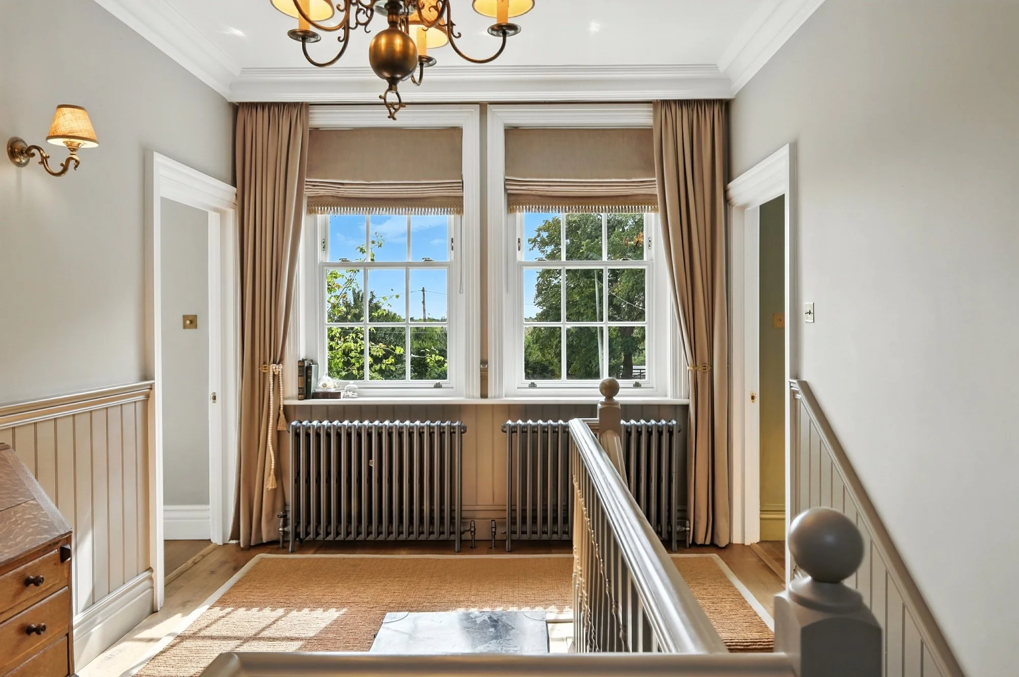 Interior view of a bright room with two large windows with beige curtains, a chandelier hanging from the ceiling, wall sconces, a radiator under the windows, and a staircase with a wooden railing.