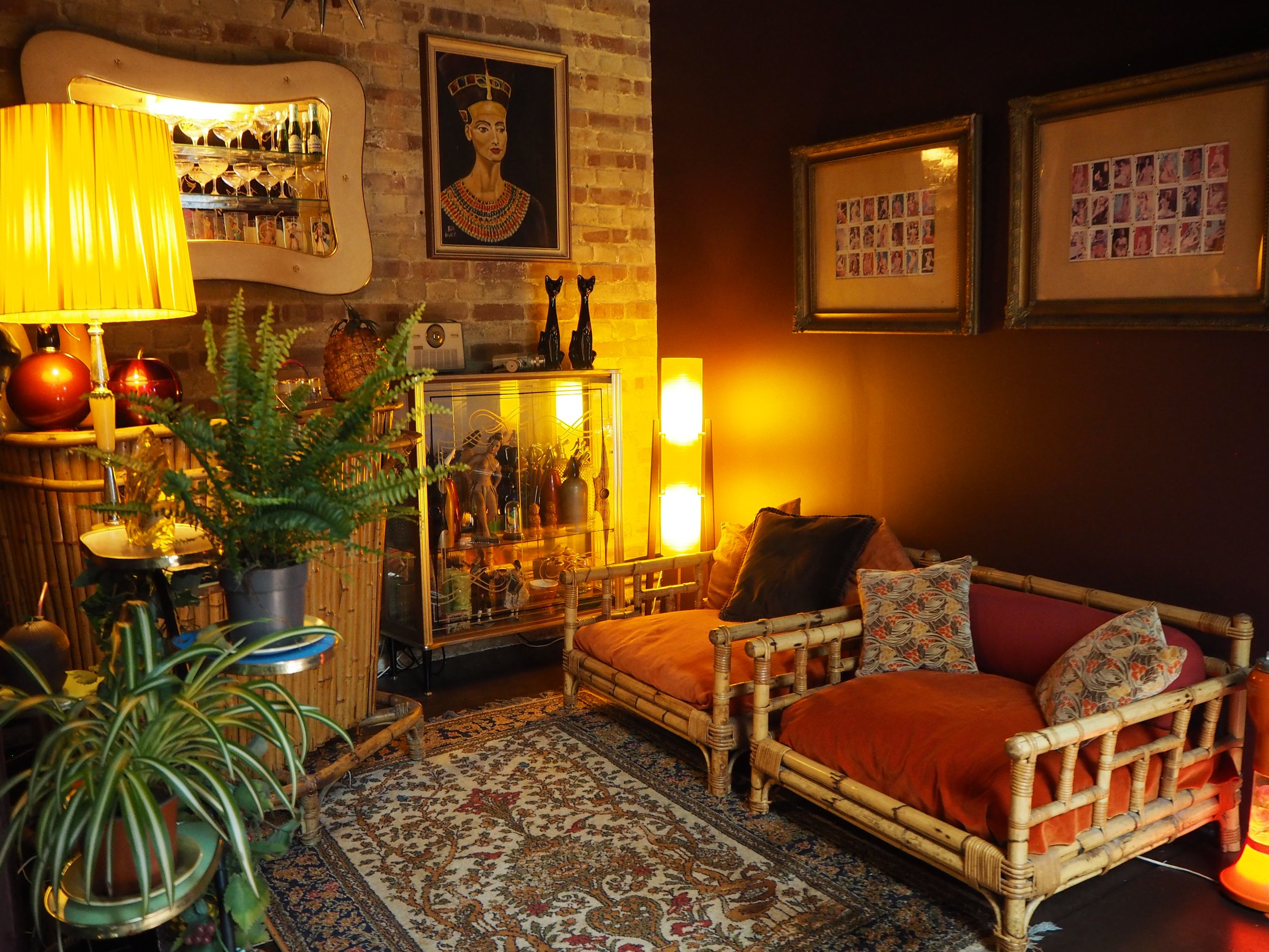 Cozy living room with bamboo furniture, plants, a glass display cabinet, framed artworks on a brick and dark wall, and warm lighting from lamps and floor lights.