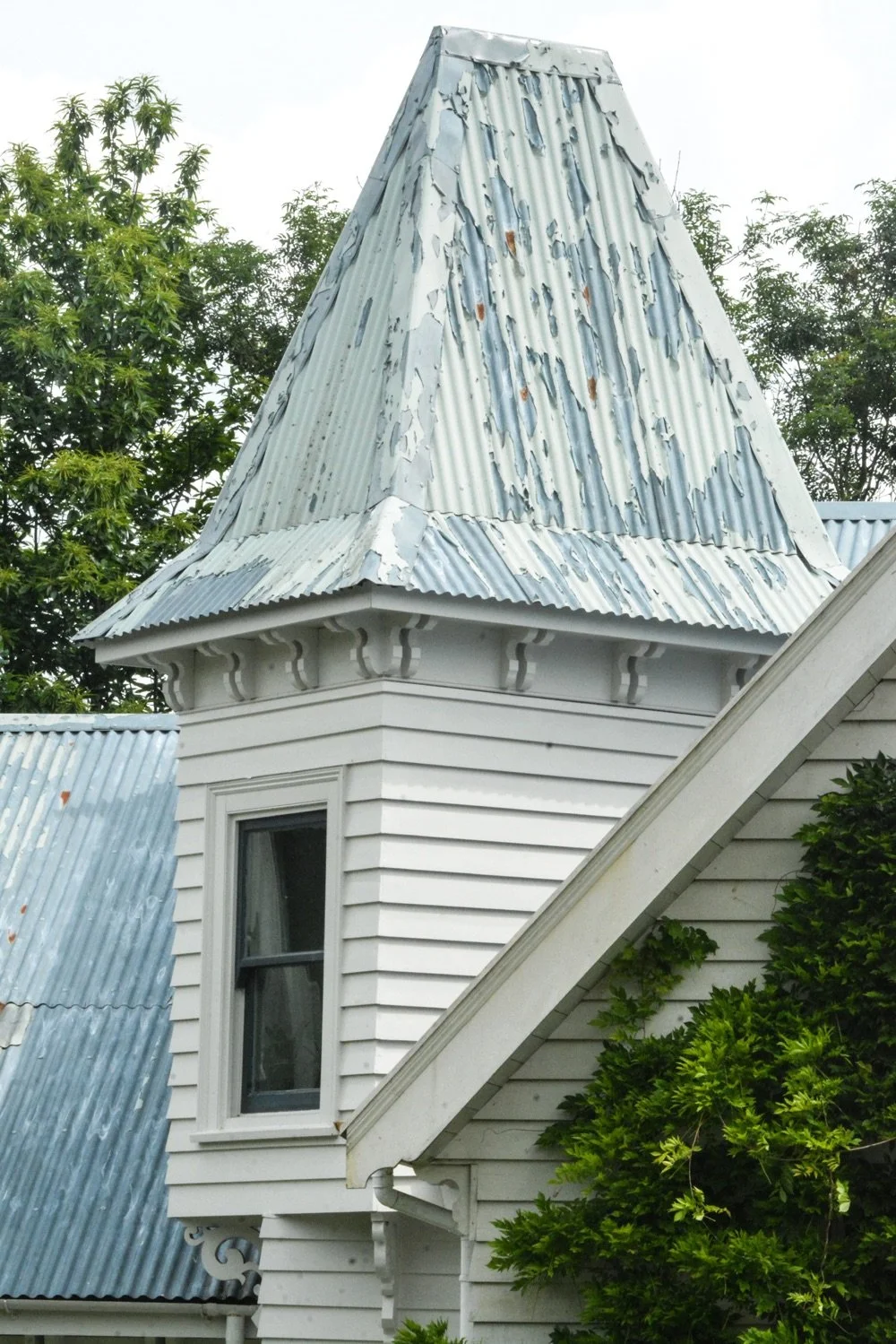 Close-up of an old, weathered metal roof on a white house with a decorative trim, surrounded by green trees.