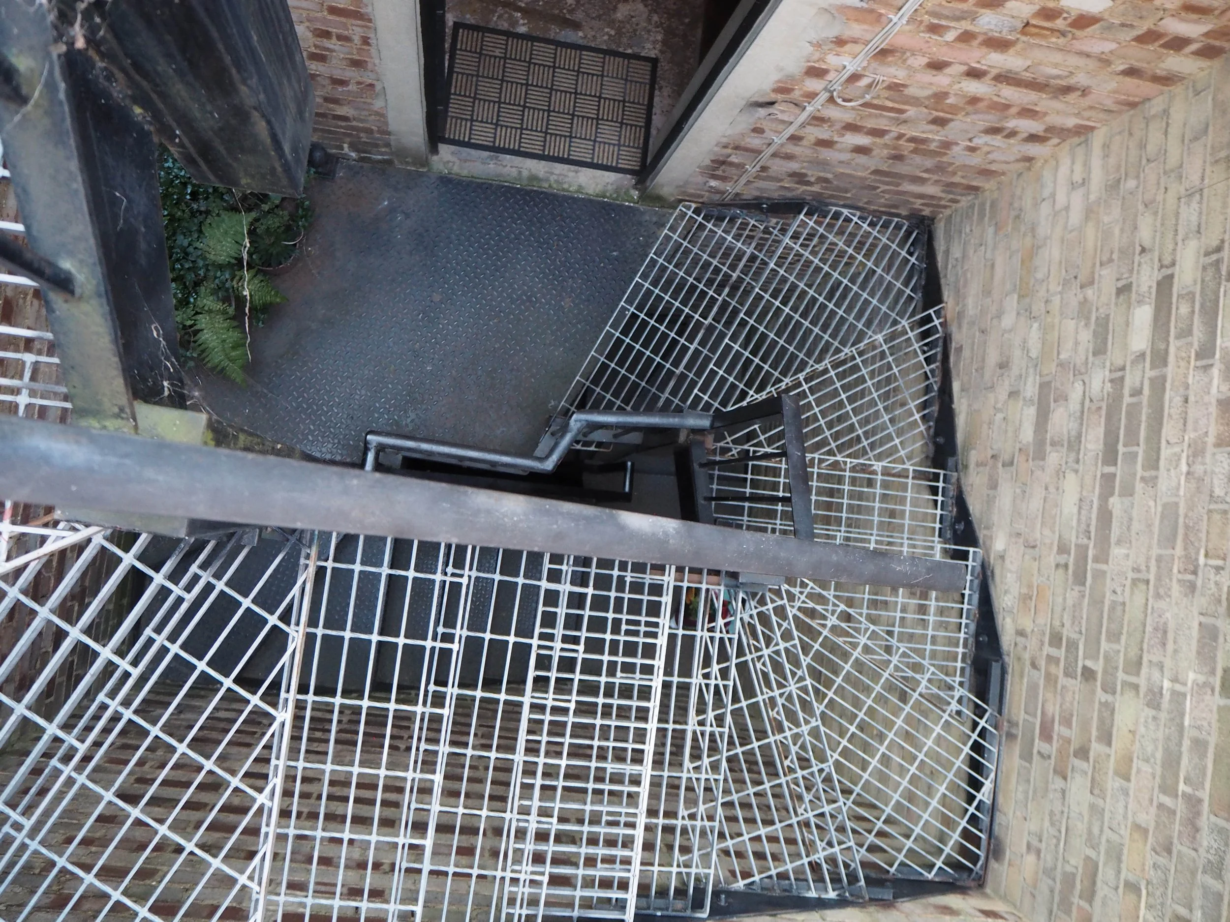 A small outdoor balcony with black metal flooring, surrounded by brick walls, and a black metal gate. Several black and white metal racks are stacked against the walls, and some ferns are growing in the corner near the black gate.