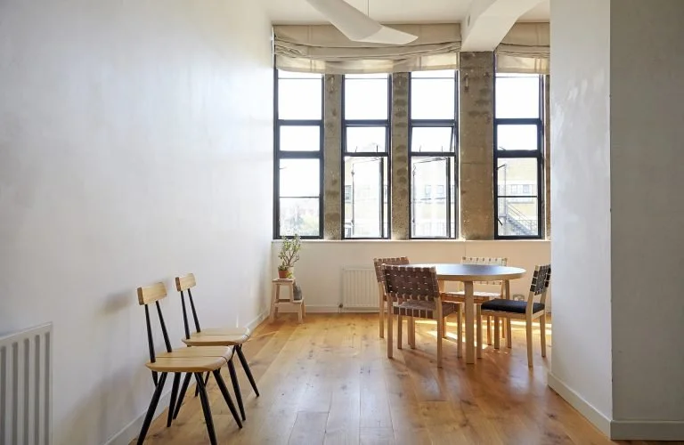 Bright dining area with large windows, wooden table, and chairs, with a potted plant in the corner.