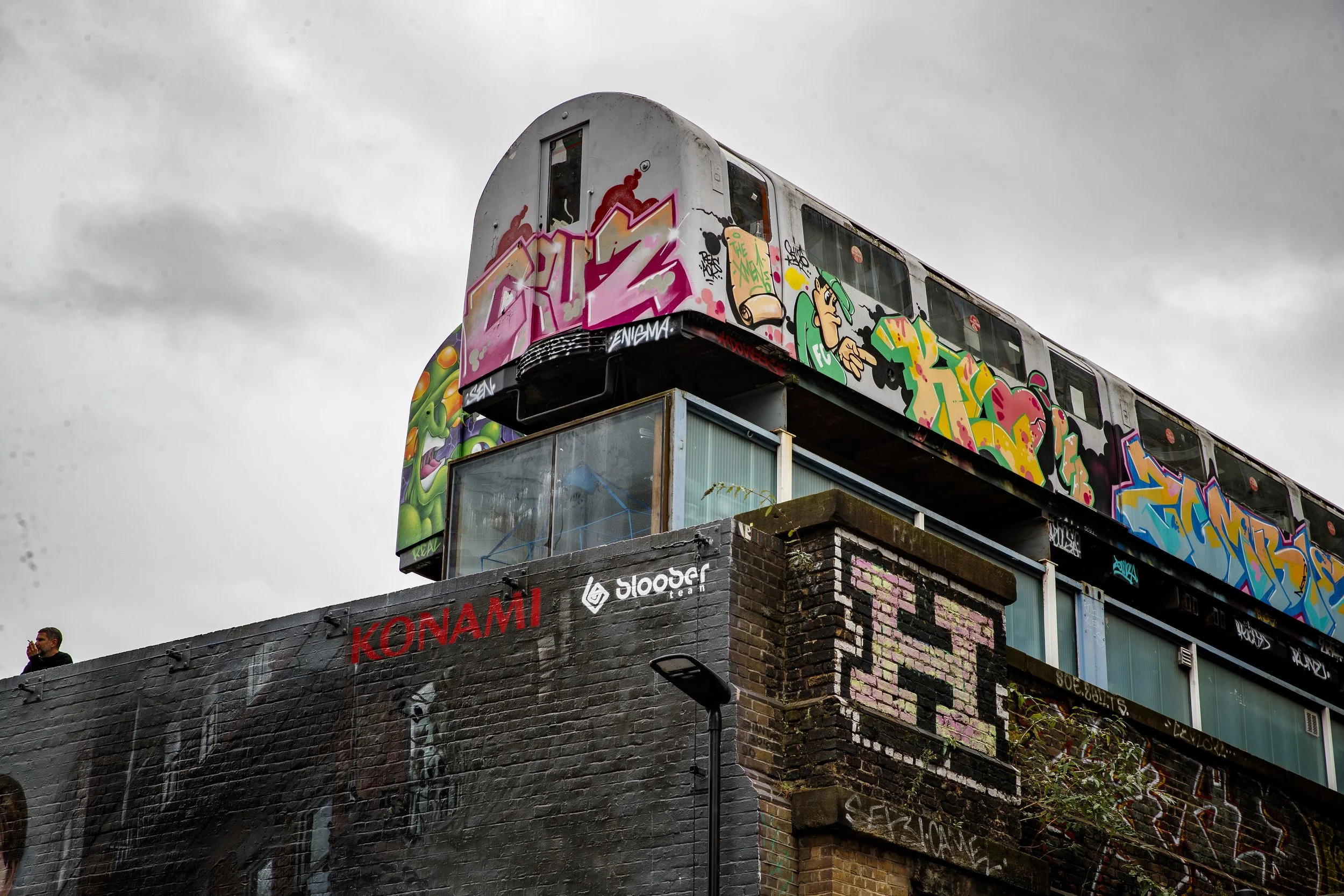 Graffiti-covered train elevated above a brick and concrete wall with shop logos, including Konami and Sloose, on a cloudy sky background.